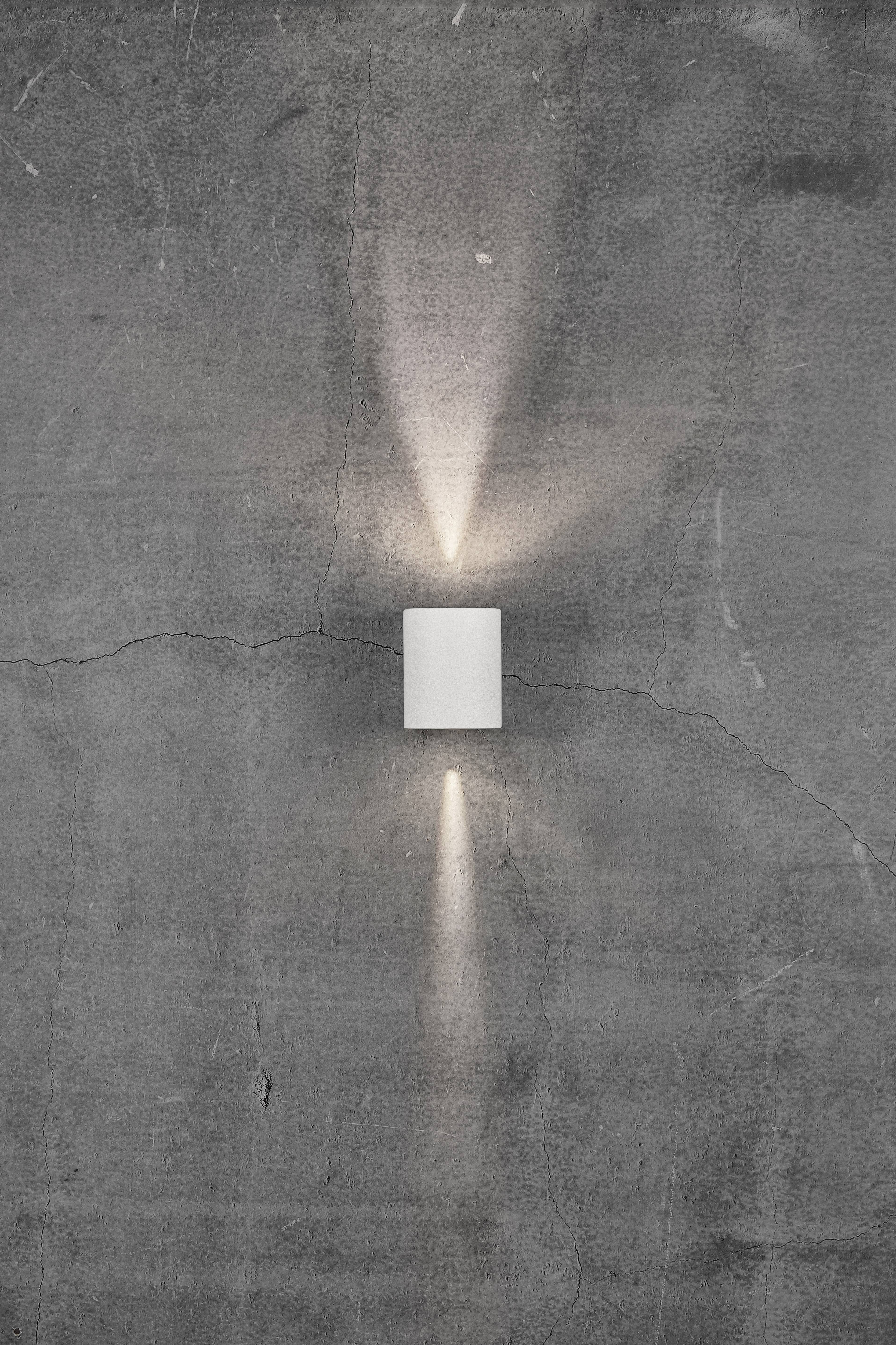 A white wall light emits light upwards and downwards onto a grey concrete wall with cracks.