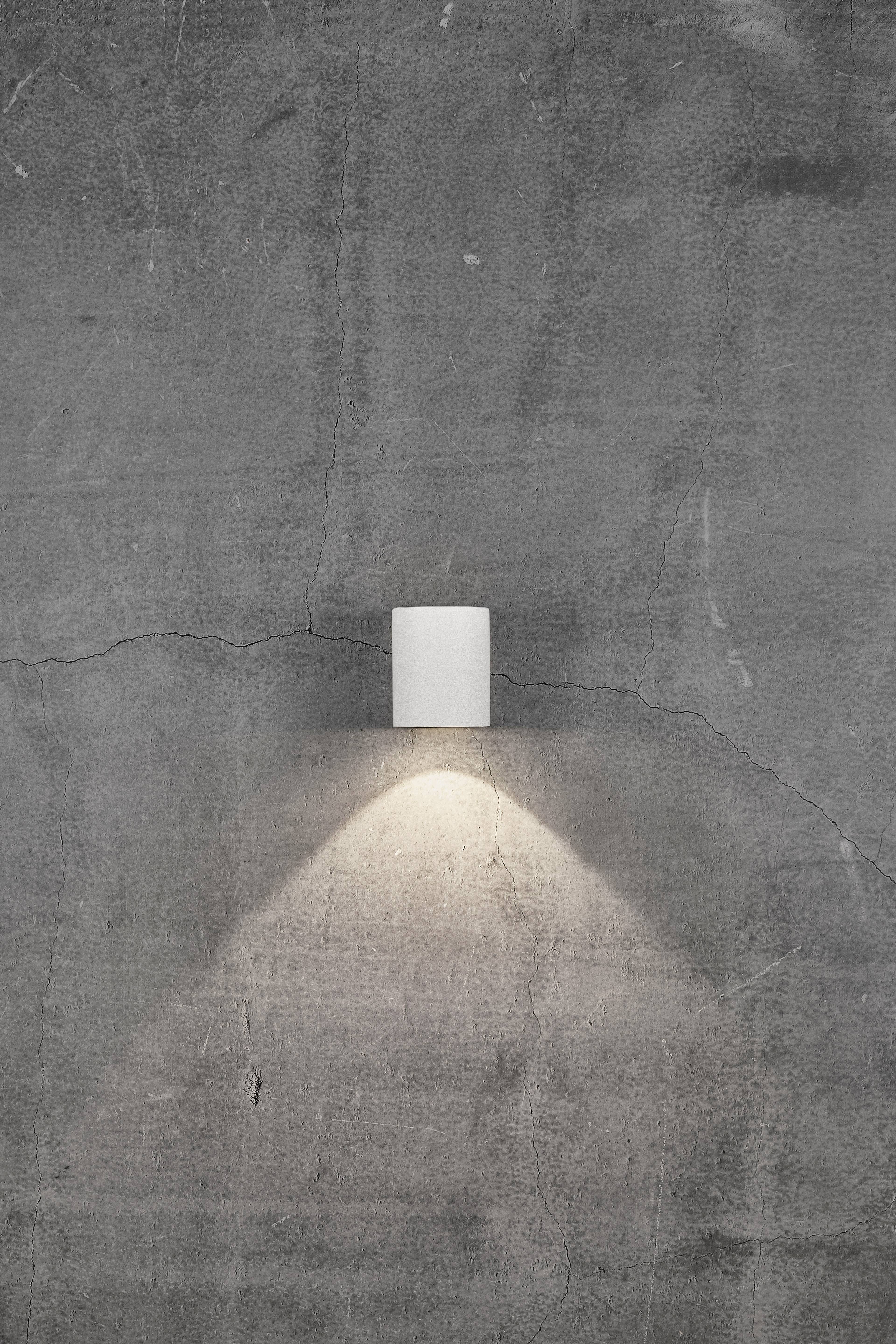 Light grey concrete wall with a small, round ceiling light casting warm light. The lamp light emphasises the wall's texture.