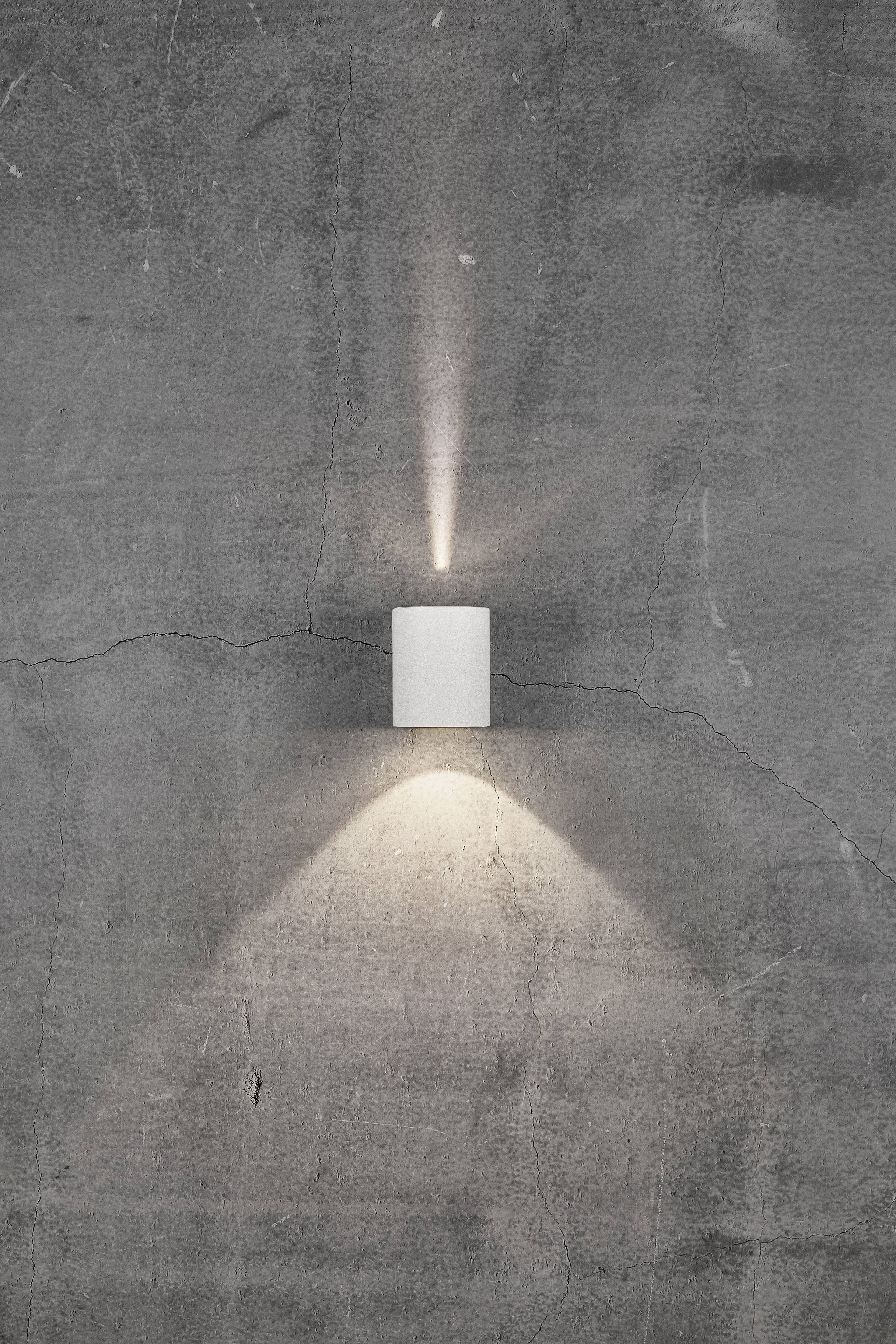 A cylindrical wall light on a concrete wall, radiating a warm, soft light upwards and downwards.