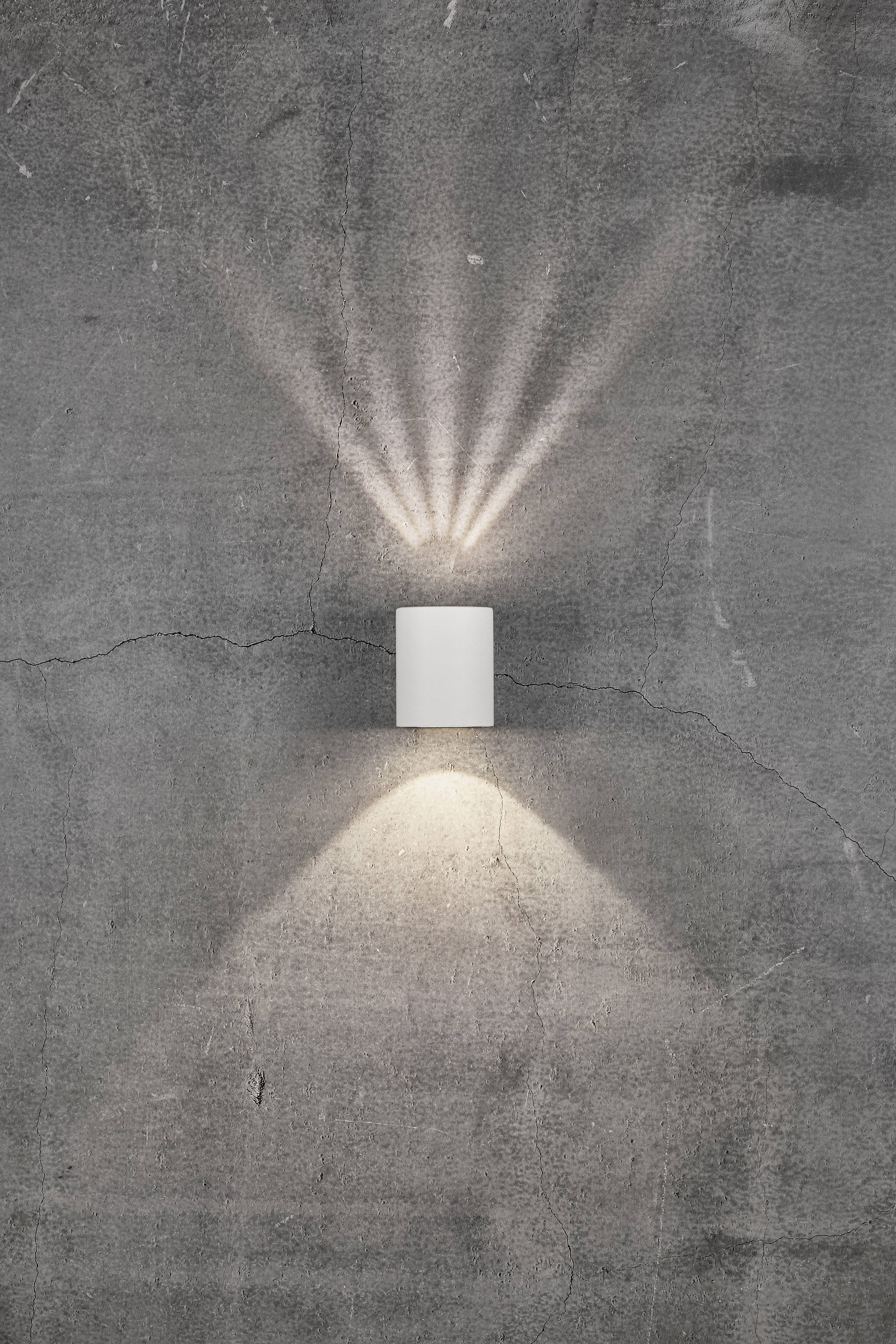 Wall light mounted on a grey concrete wall, casting light rays upwards and downwards, creating patterns with triangular shapes.
