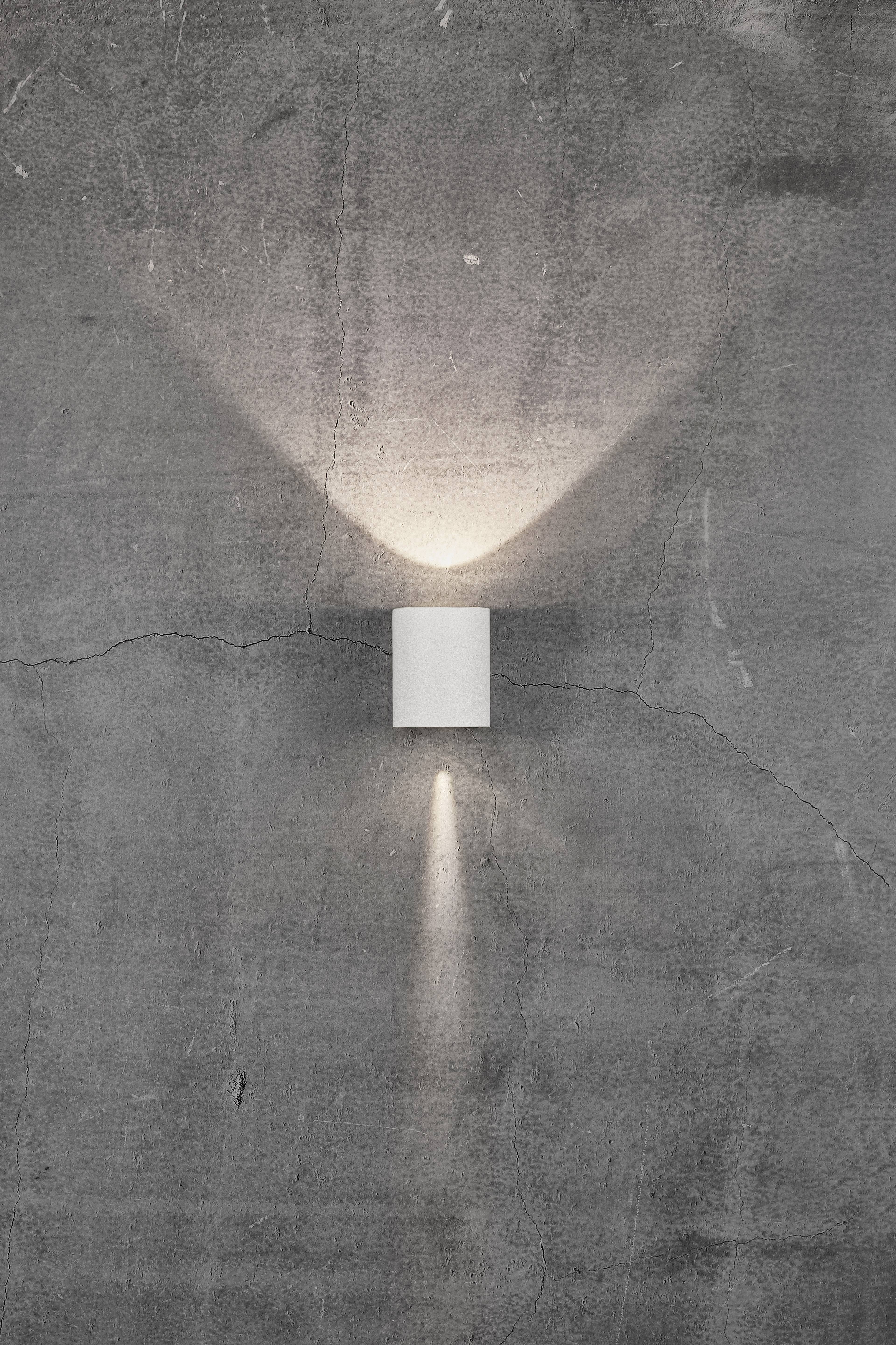 Grey concrete wall with a cylindrical wall light. Light radiates upwards and downwards, creating a V-shaped pattern.