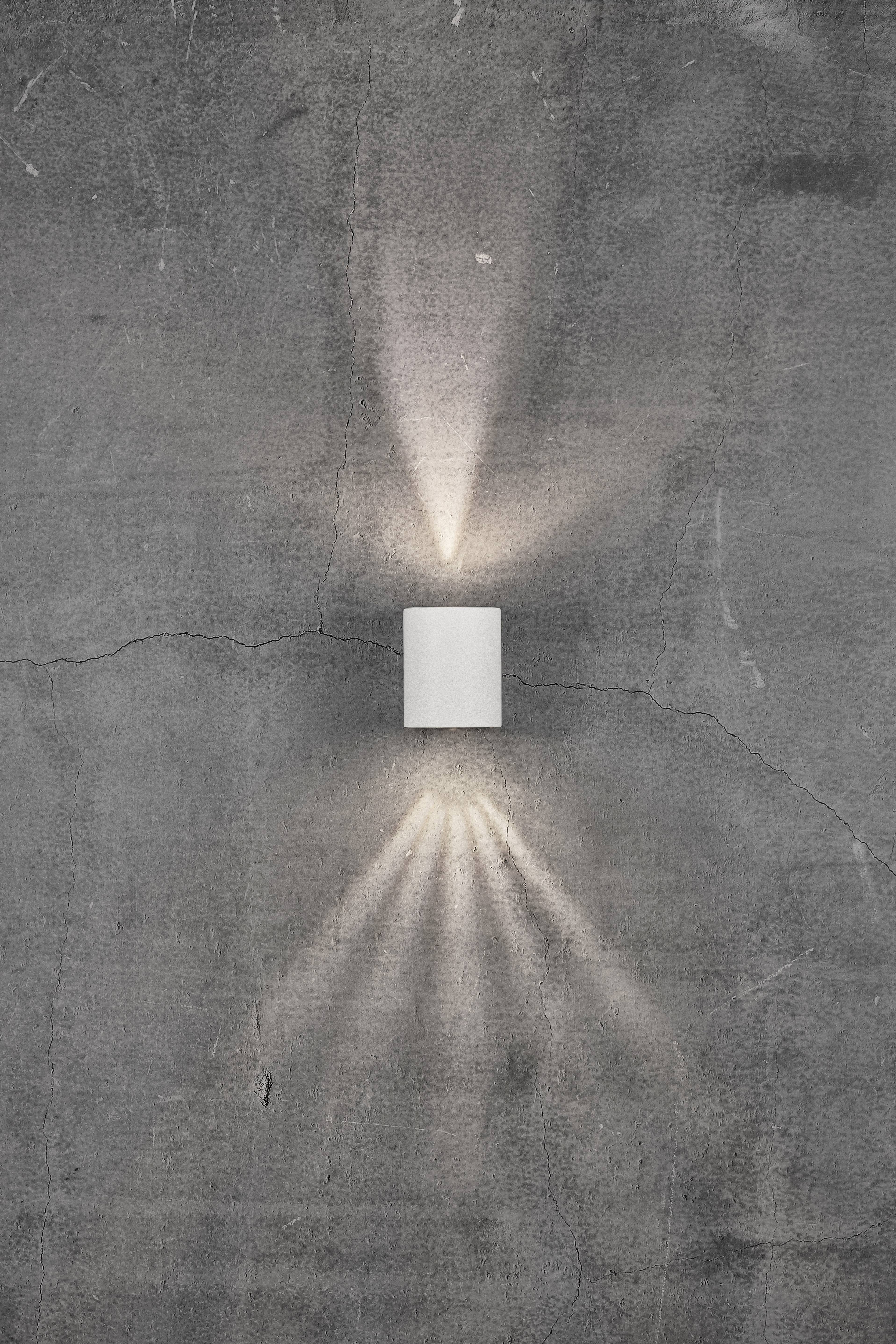 A white wall light on a grey, cracked wall, casting light upwards and downwards and creating a pattern of light and shadow.