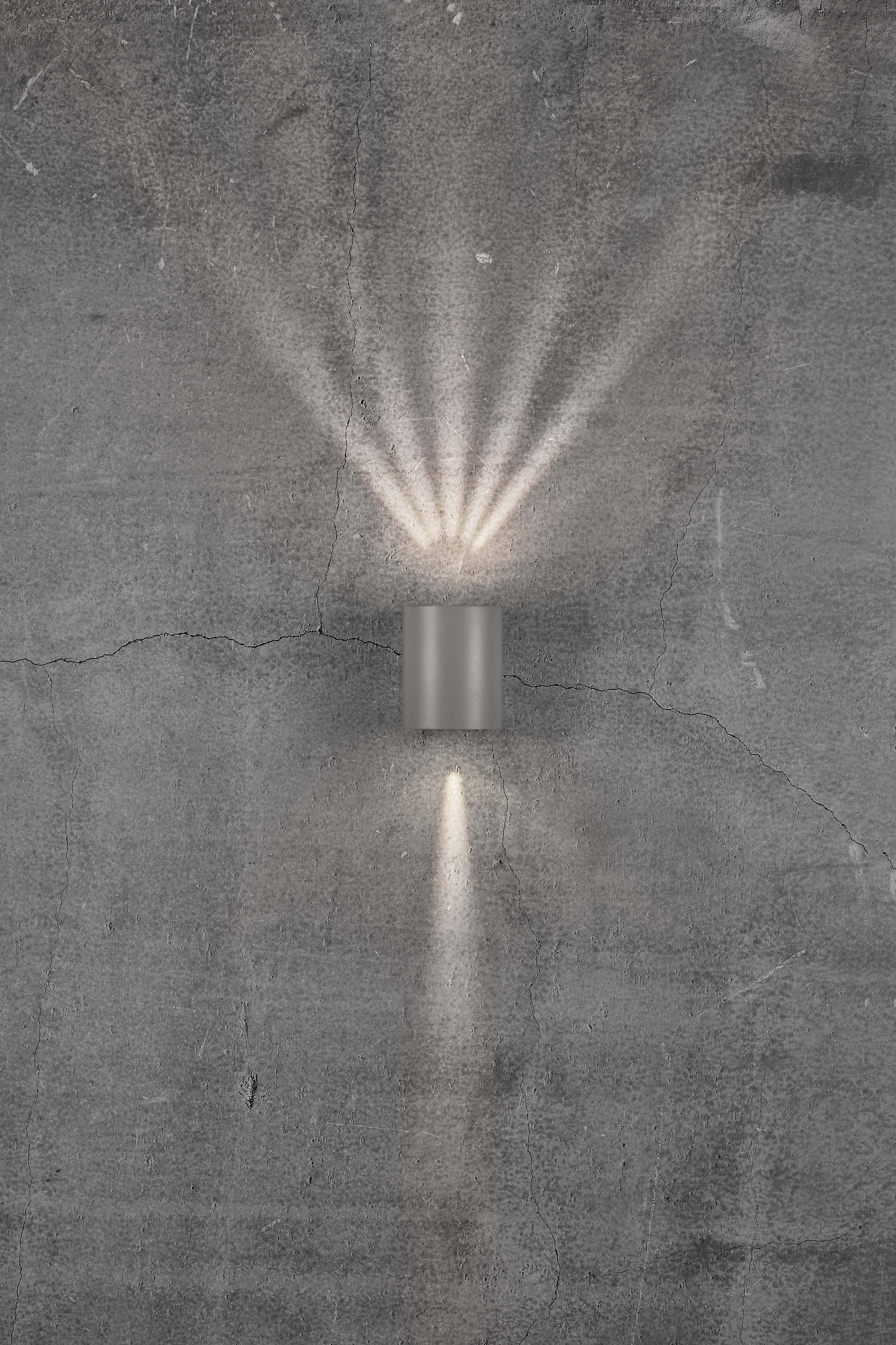 Grey wall lamp radiates warm light upwards and downwards onto a rough, grey concrete wall.