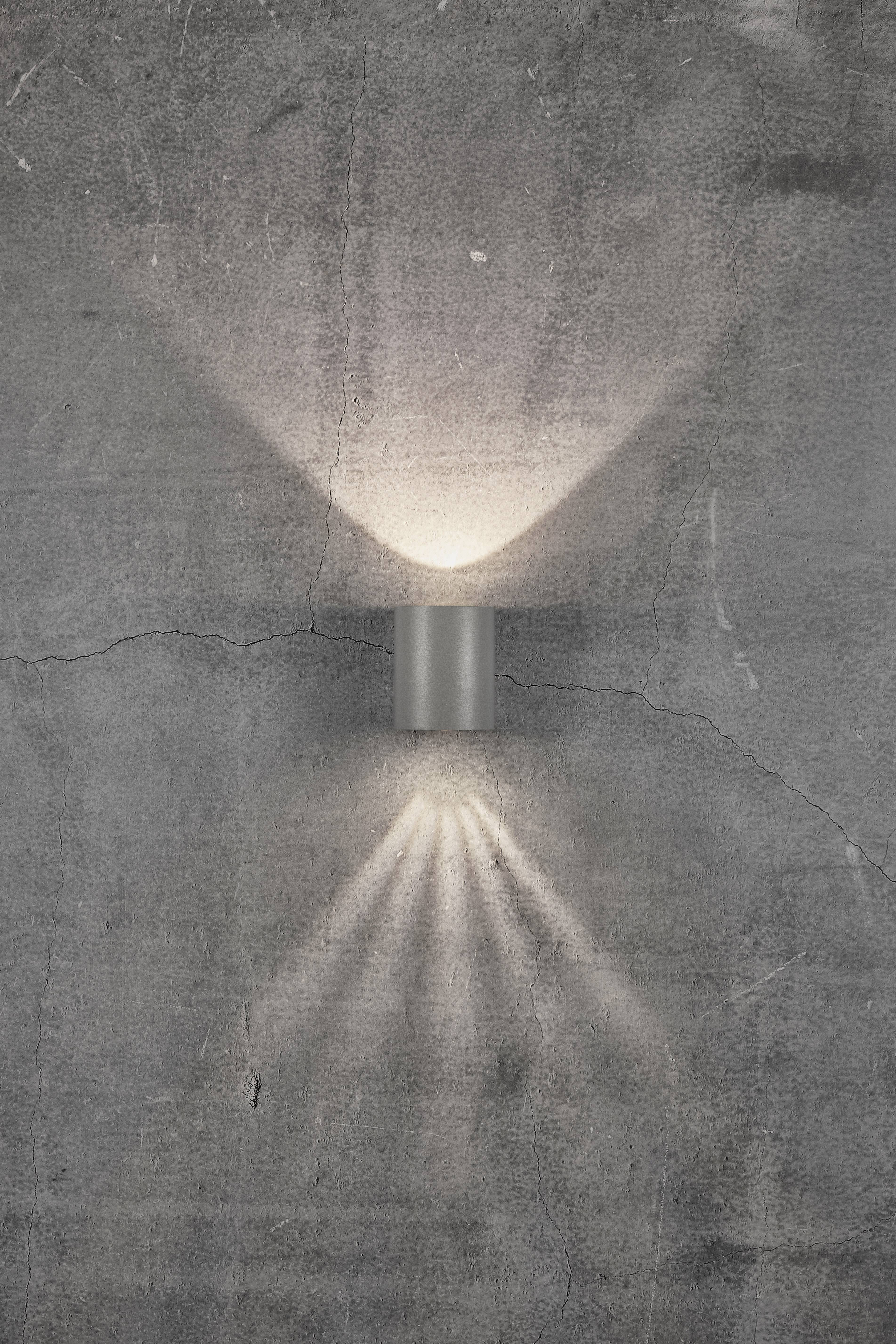Wall light made of metal casts symmetrical light rays upwards and downwards onto a grey concrete wall.