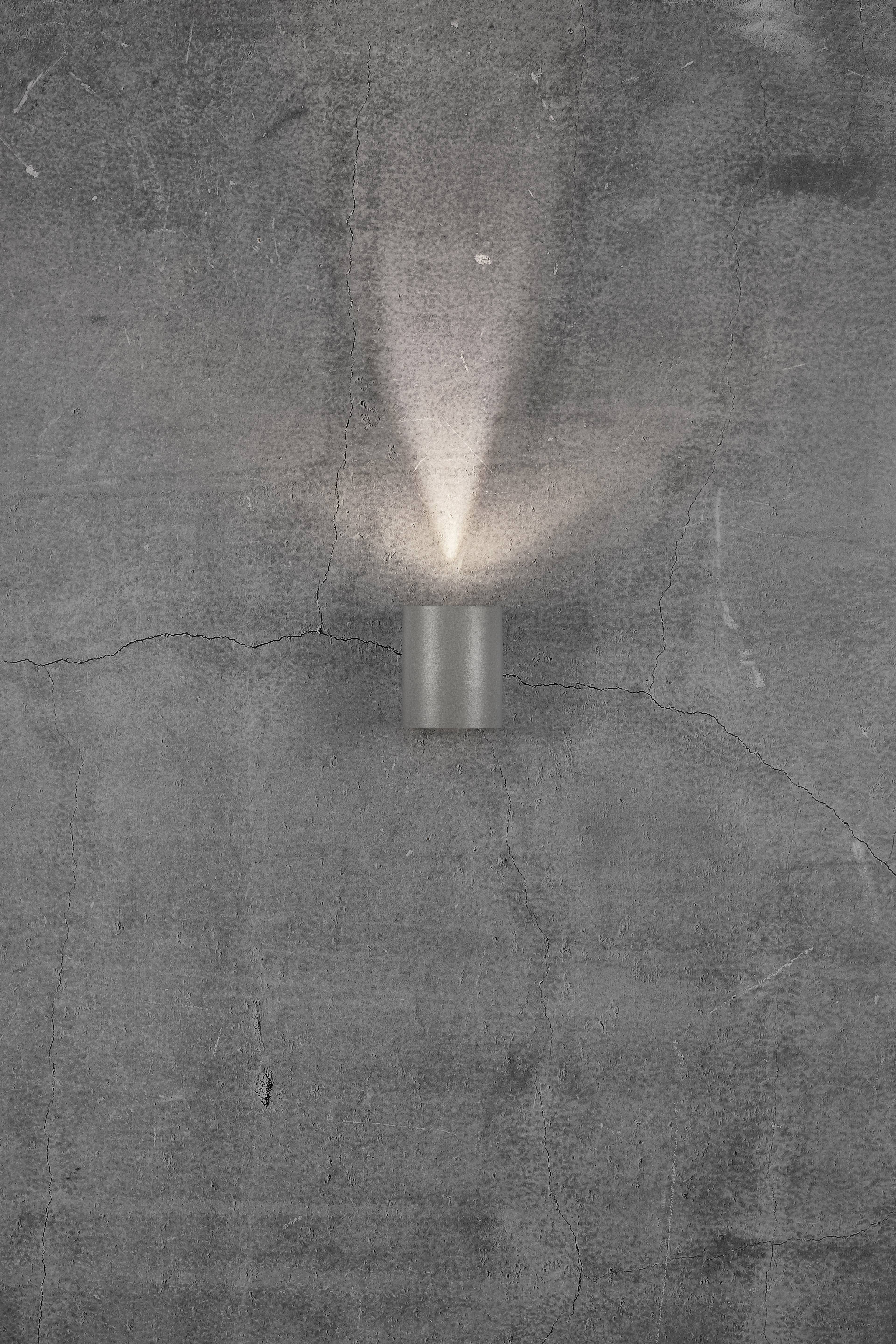 A grey concrete wall with a modern, cylindrical wall light that shines upwards against the wall, creating a light cone.