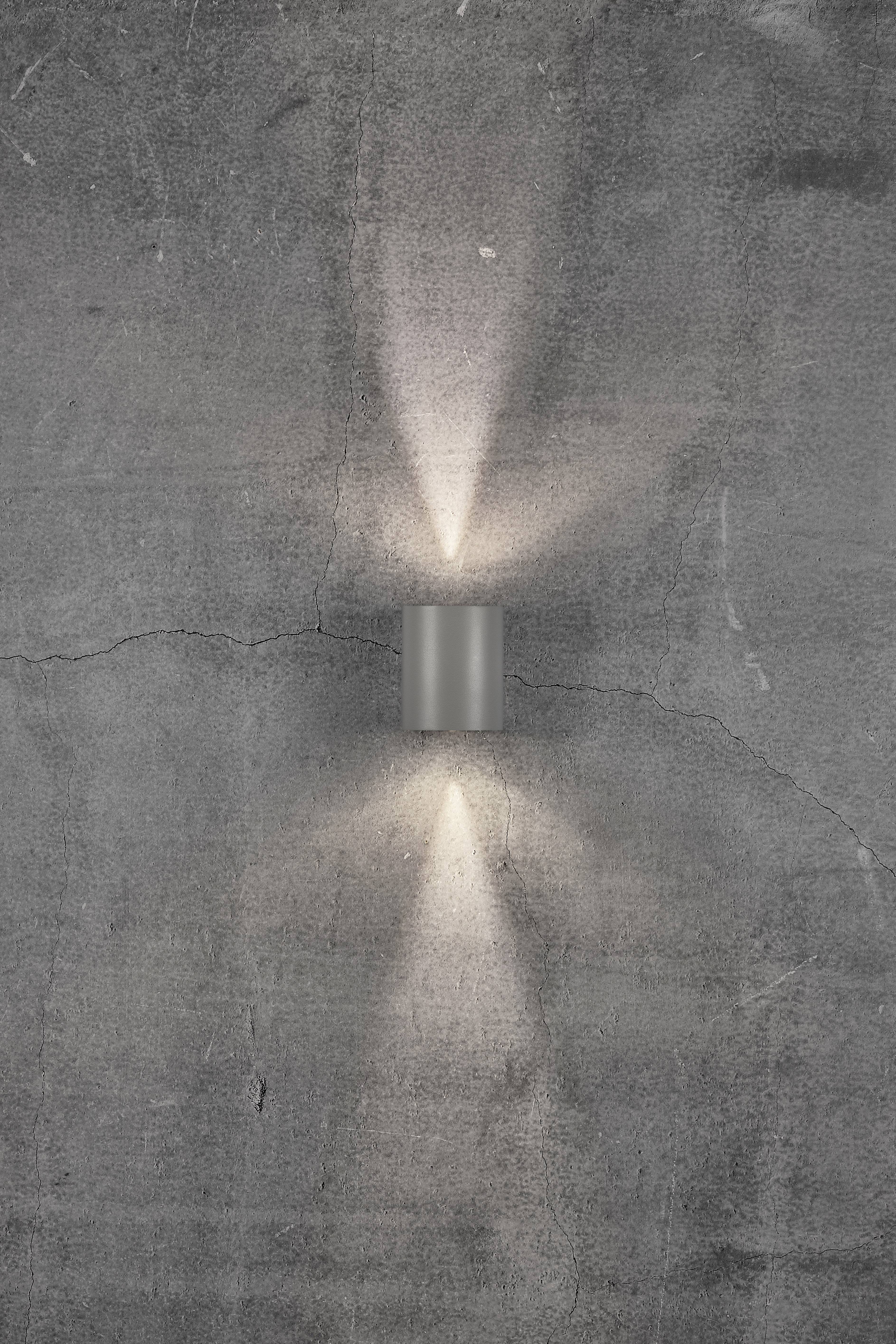 A wall light in a cylindrical shape casts warm light upwards and downwards onto a grey concrete wall with fine cracks.