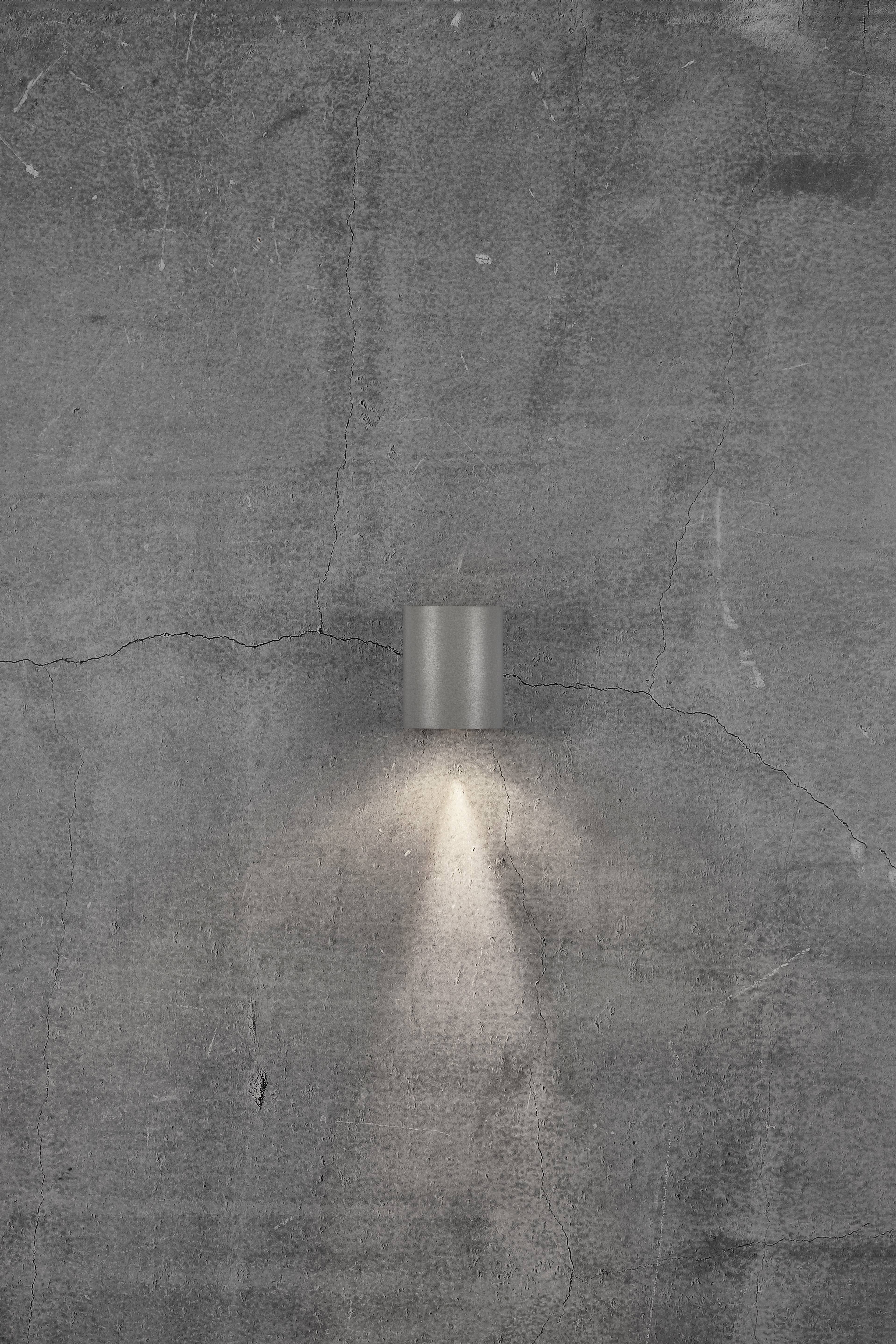 A wall light illuminates in a cone onto a grey, structural concrete wall, creating a soft light pattern downwards.