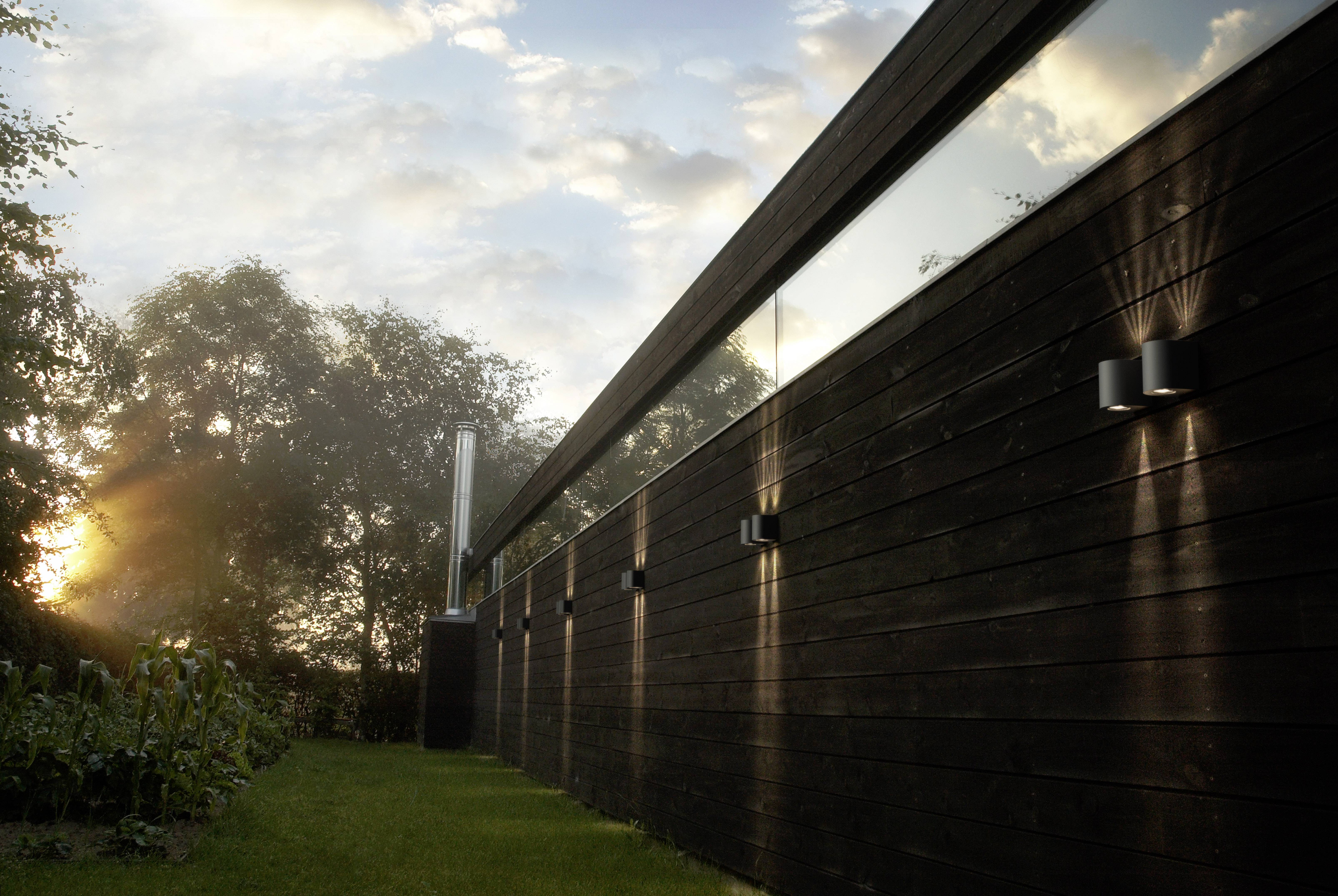A modern, elongated building with a wooden facade and narrow windows, illuminated by exterior lights, at sunset in a natural setting.