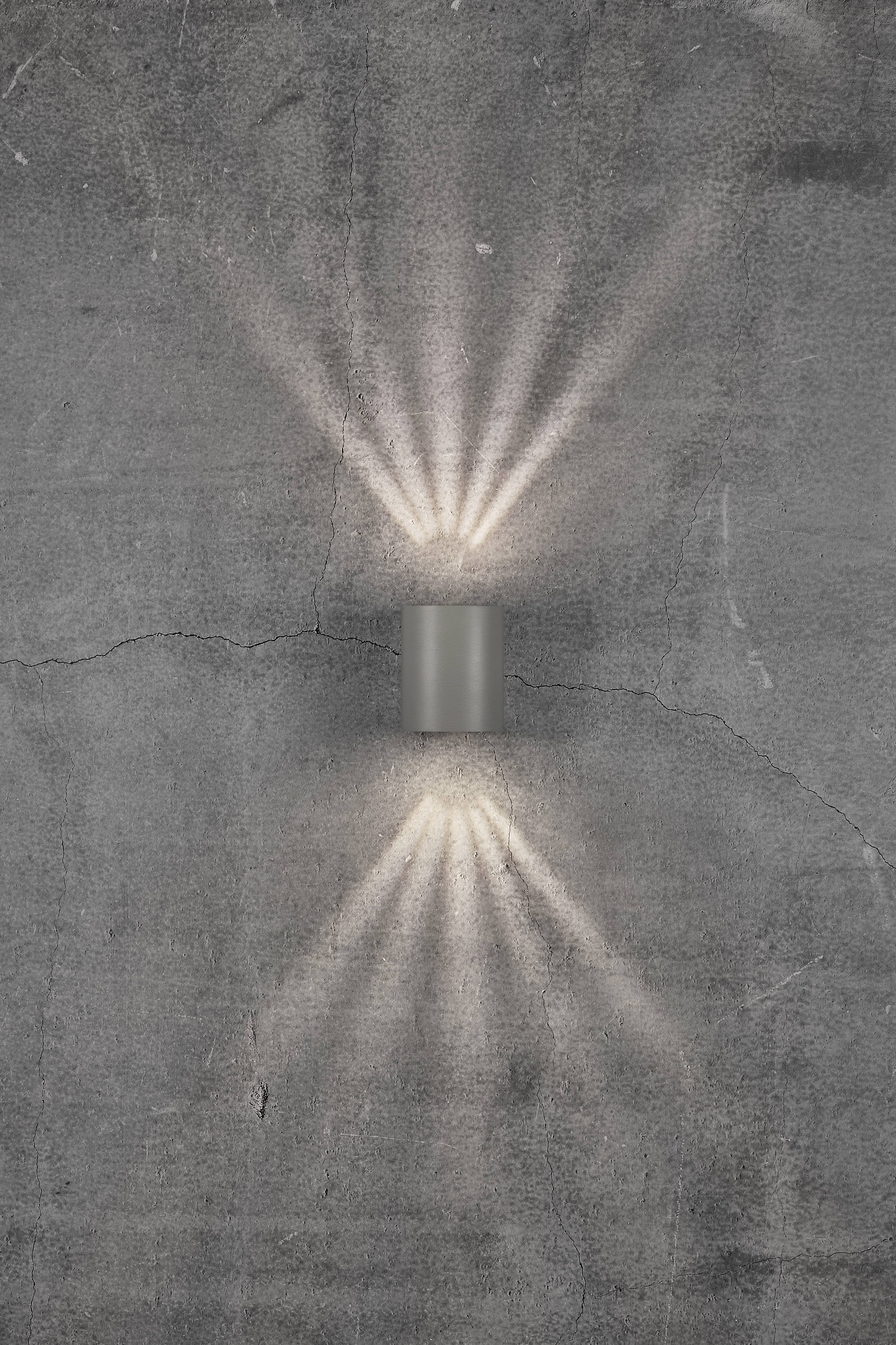 A grey wall light emits light rays upwards and downwards onto a rough, grey concrete wall.