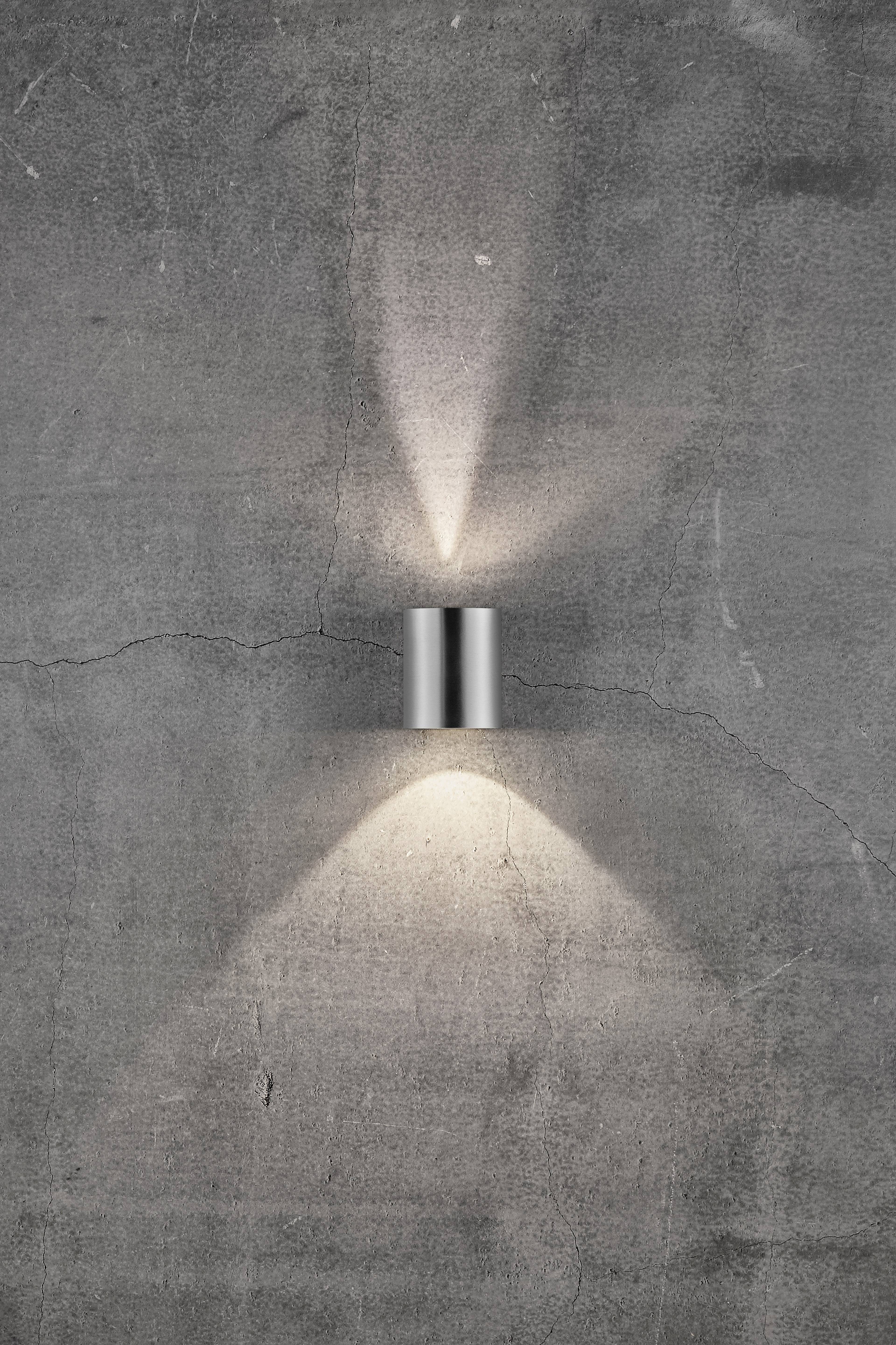 Silver wall light with two light beams, one pointing upwards and one pointing downwards, mounted on a grey concrete wall.