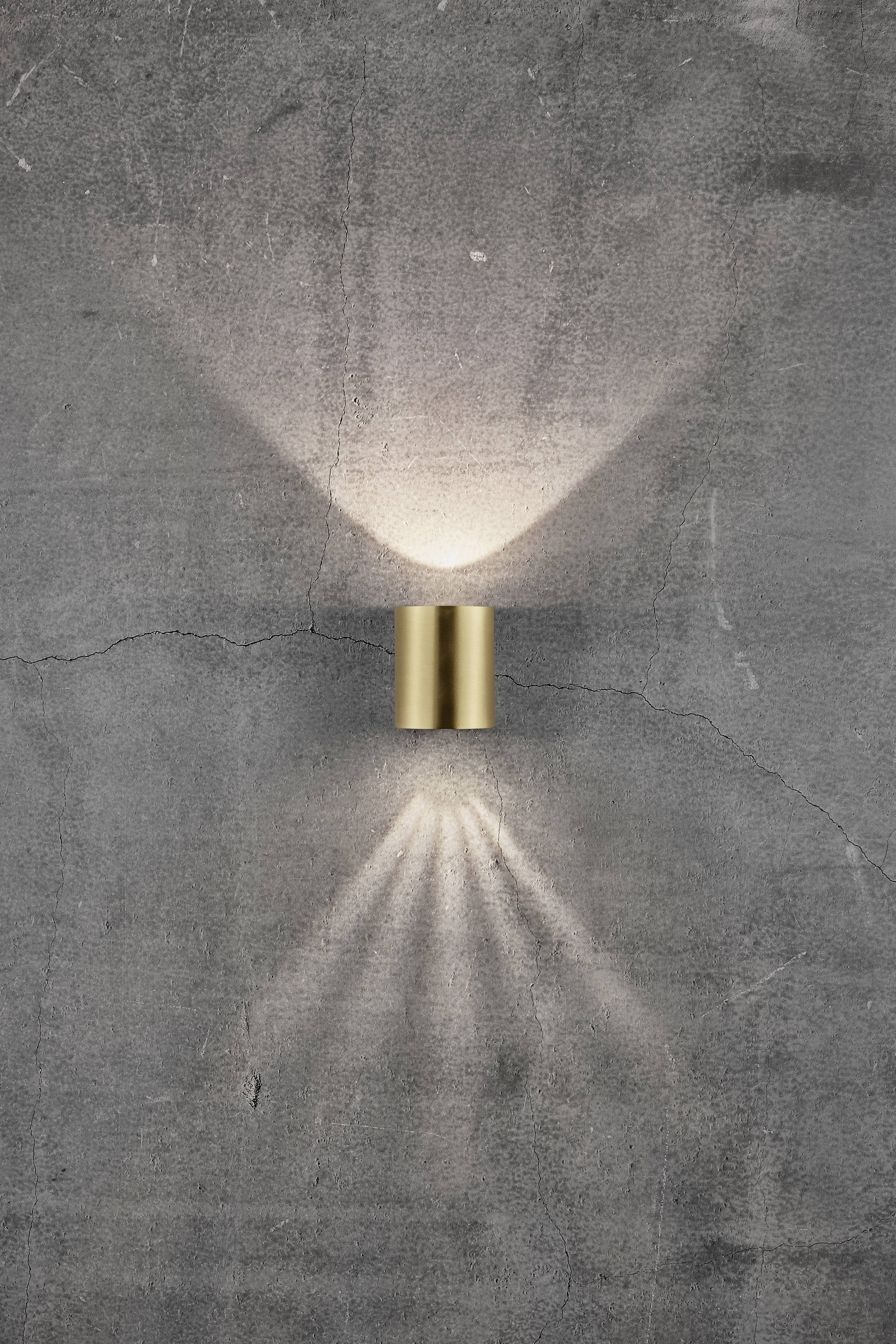 Wall light mounted on a concrete wall, projecting light rays upwards and downwards. Modern design with brushed brass.