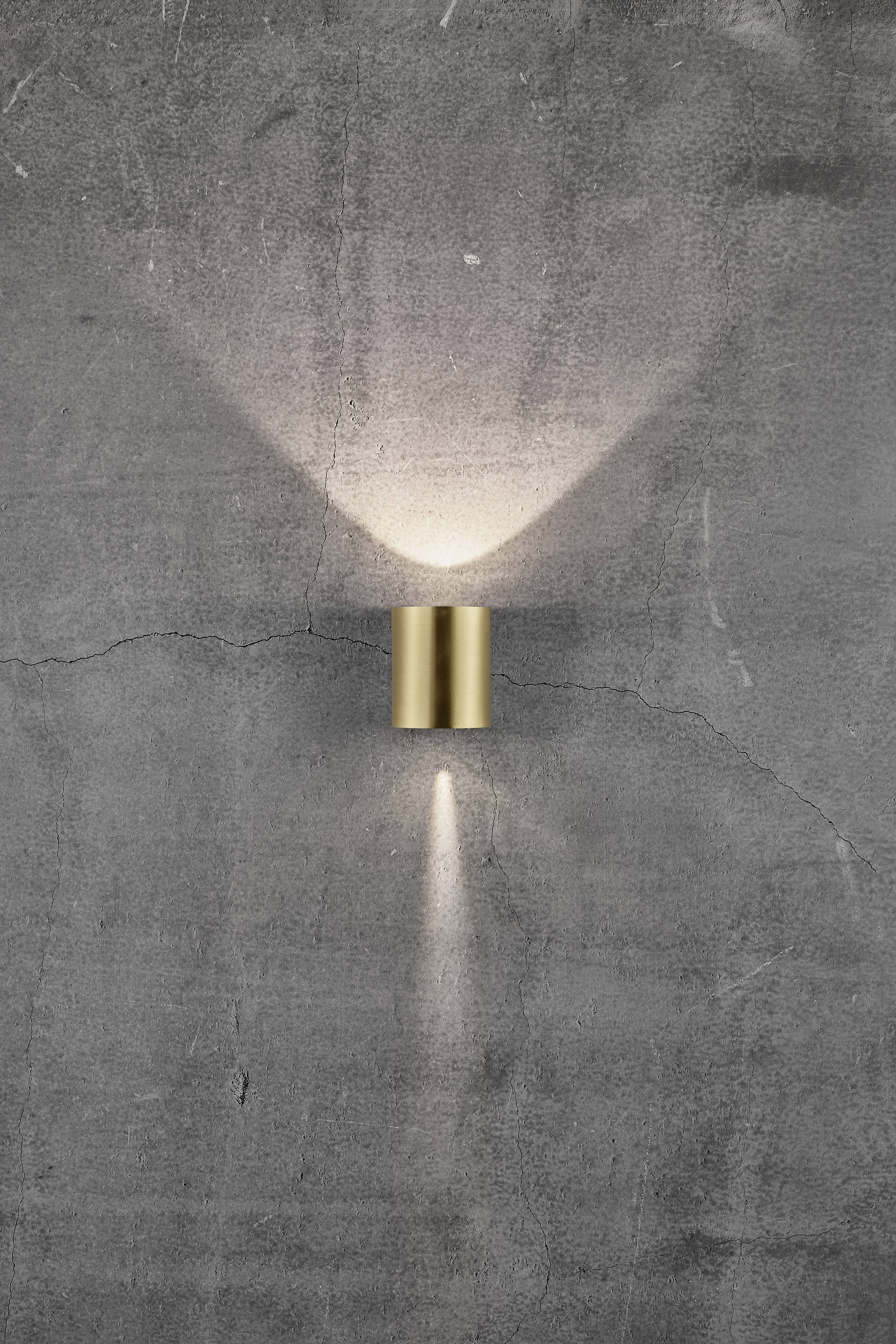Wall light made of brass on a grey, textured wall. The light radiates upwards and downwards, creating accents on the wall.