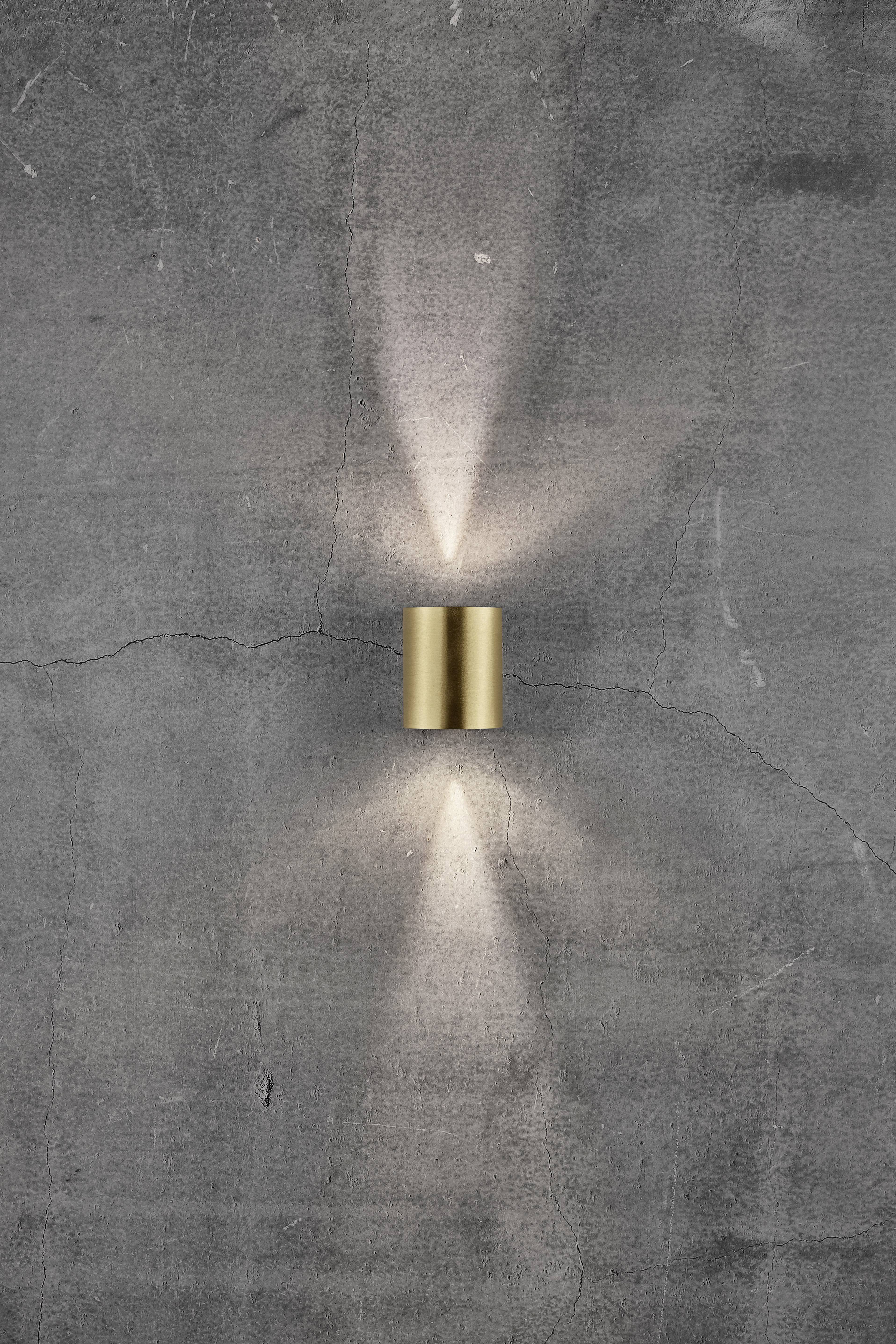 Wall light made of brass casts warm light upwards and downwards onto a grey concrete wall, creating a stylish light pattern.