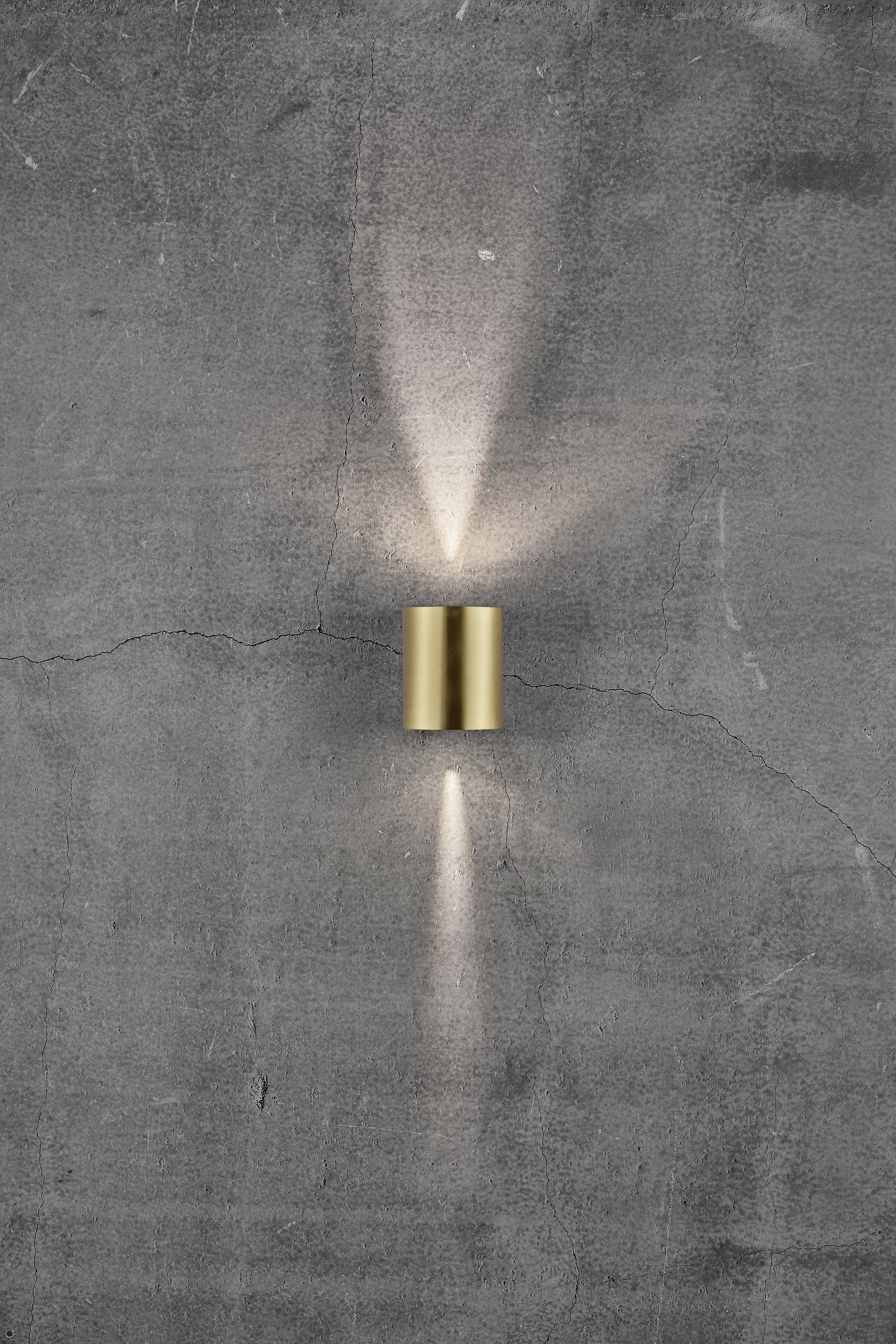 A cylindrical, golden wall light on a grey, slightly cracked surface, emitting light rays upwards and downwards.