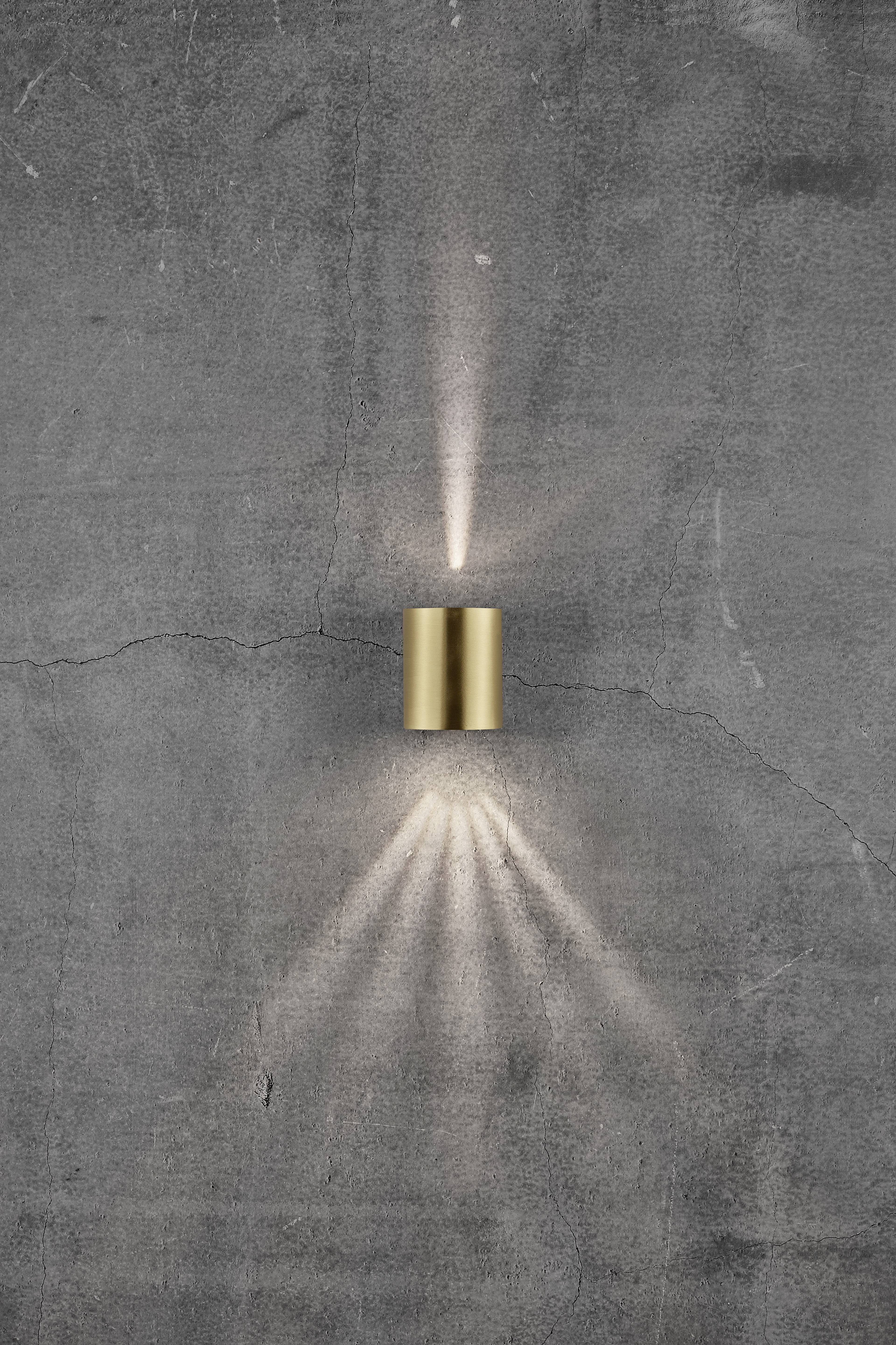 A cylindrical, golden wall element casts light rays upwards and downwards onto a grey, textured wall.