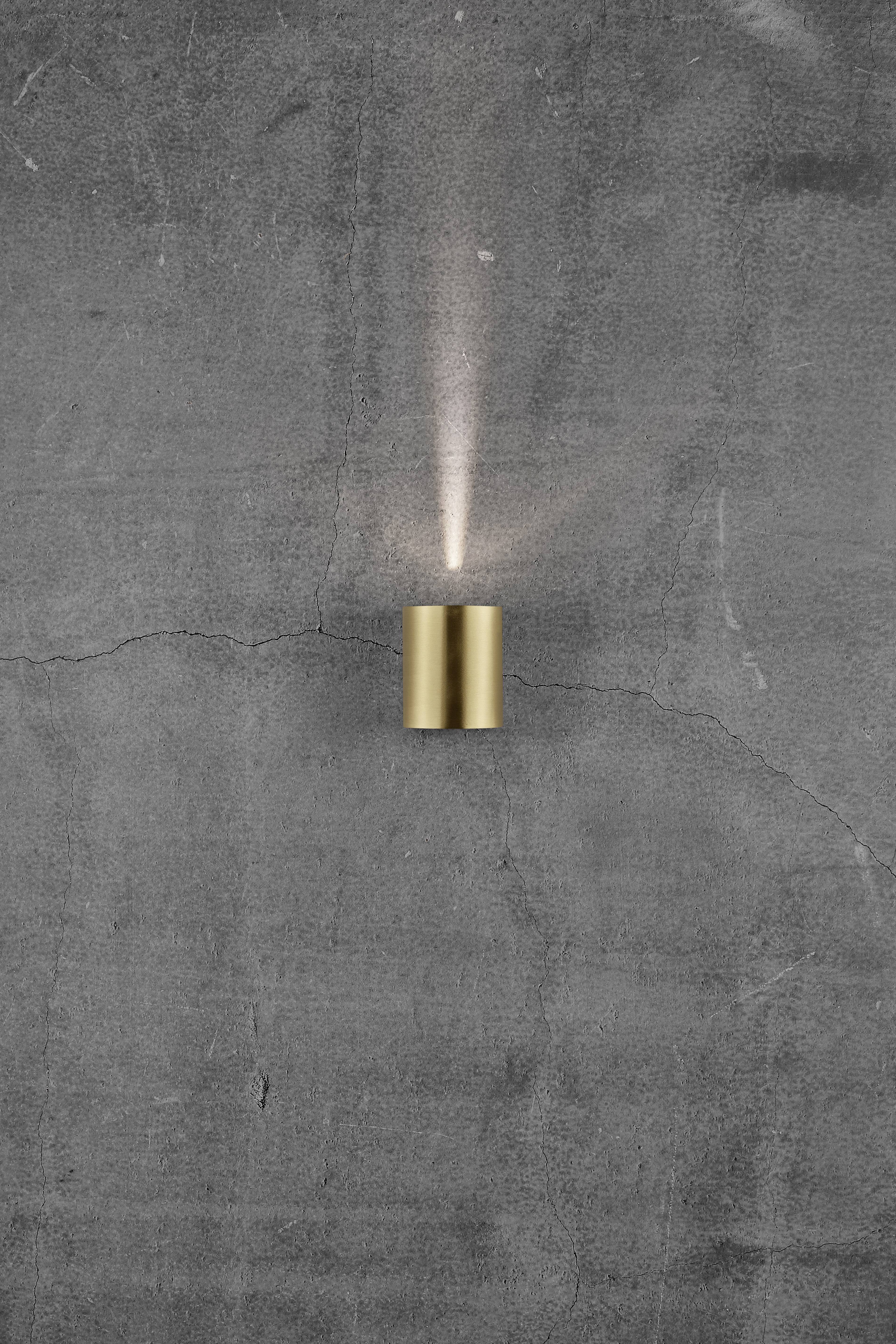 Wall with a grey concrete surface, to which a small, stylised, golden wall light is affixed, casting a soft upward glow.