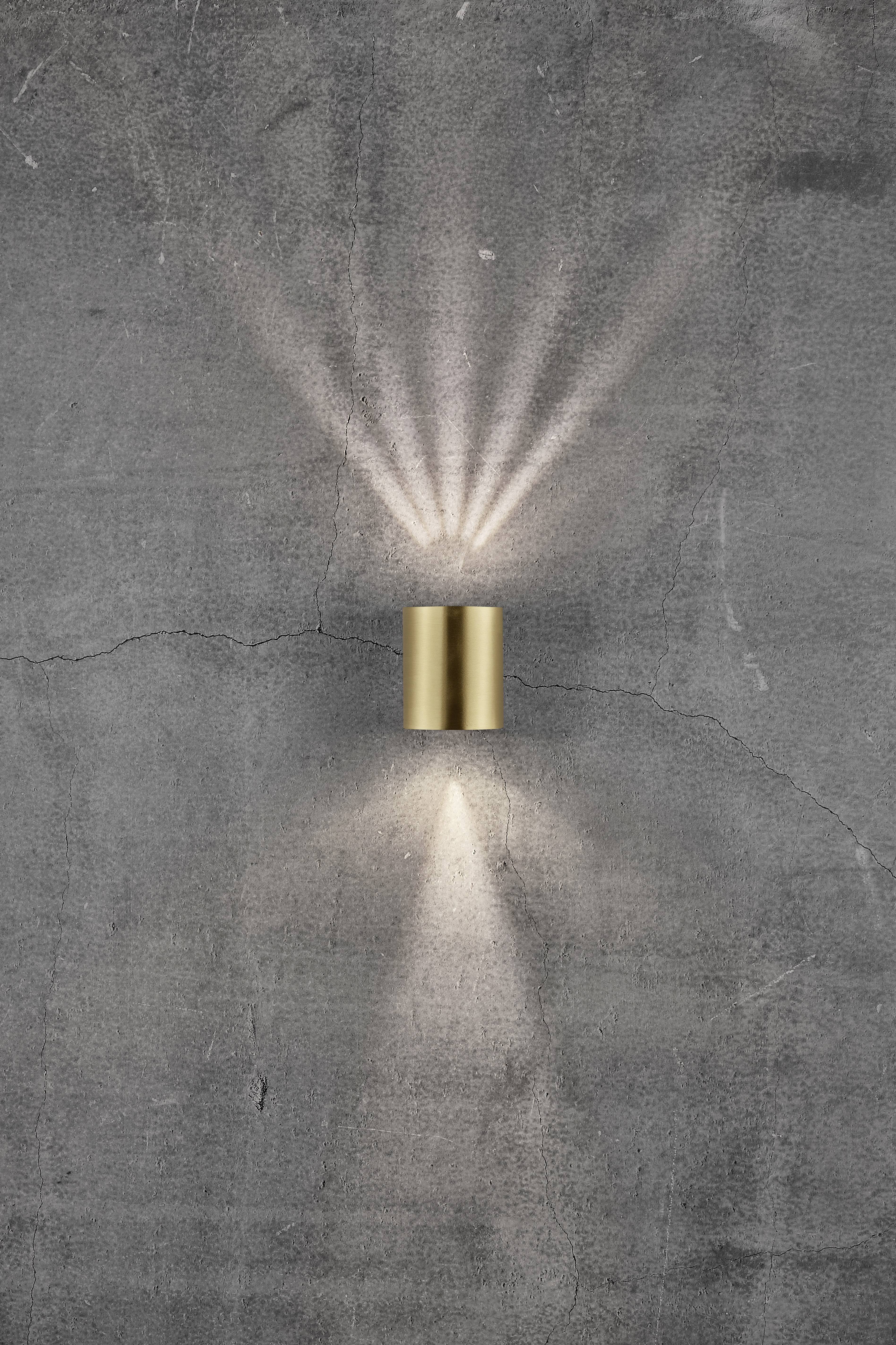 Brass wall light mounted on a grey, textured wall. Light radiates upwards and downwards, creating decorative patterns on the surface.