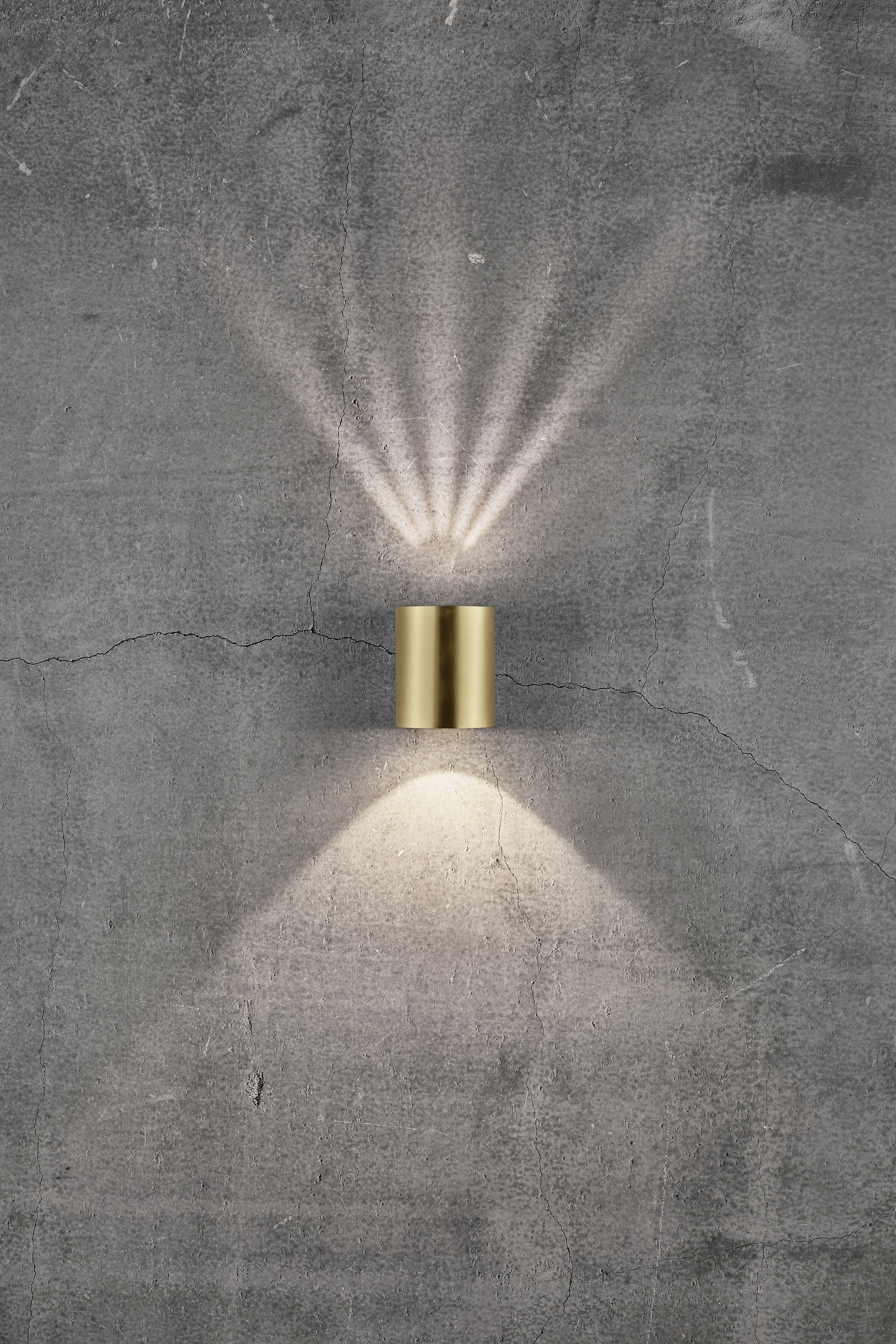 A golden wall light casts light upwards and downwards onto a grey, textured wall, creating patterned light rays.