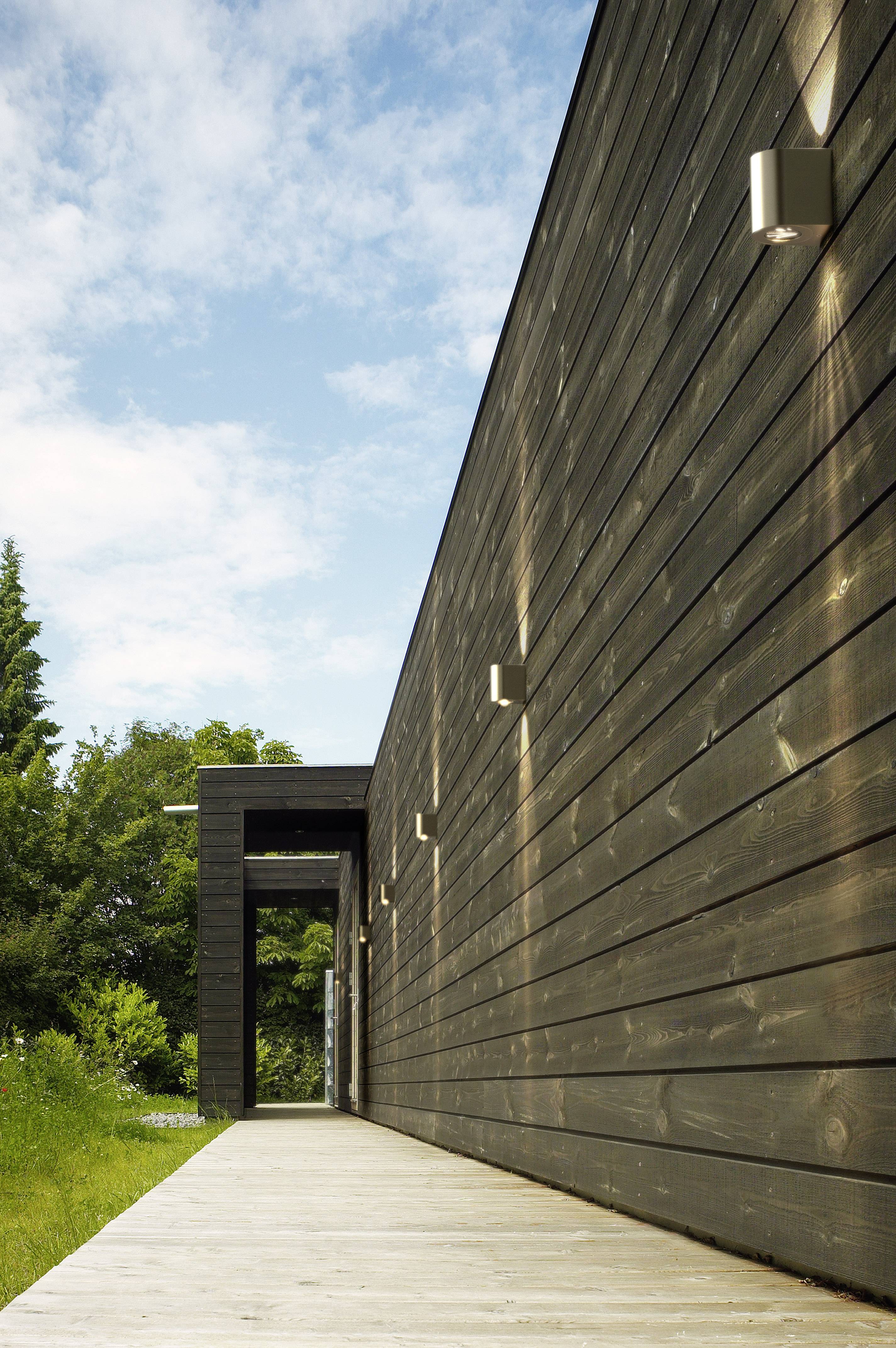 Modern building with a timber façade and bright external lighting, flanked by a wooden walkway. In the background, a blue sky is visible.