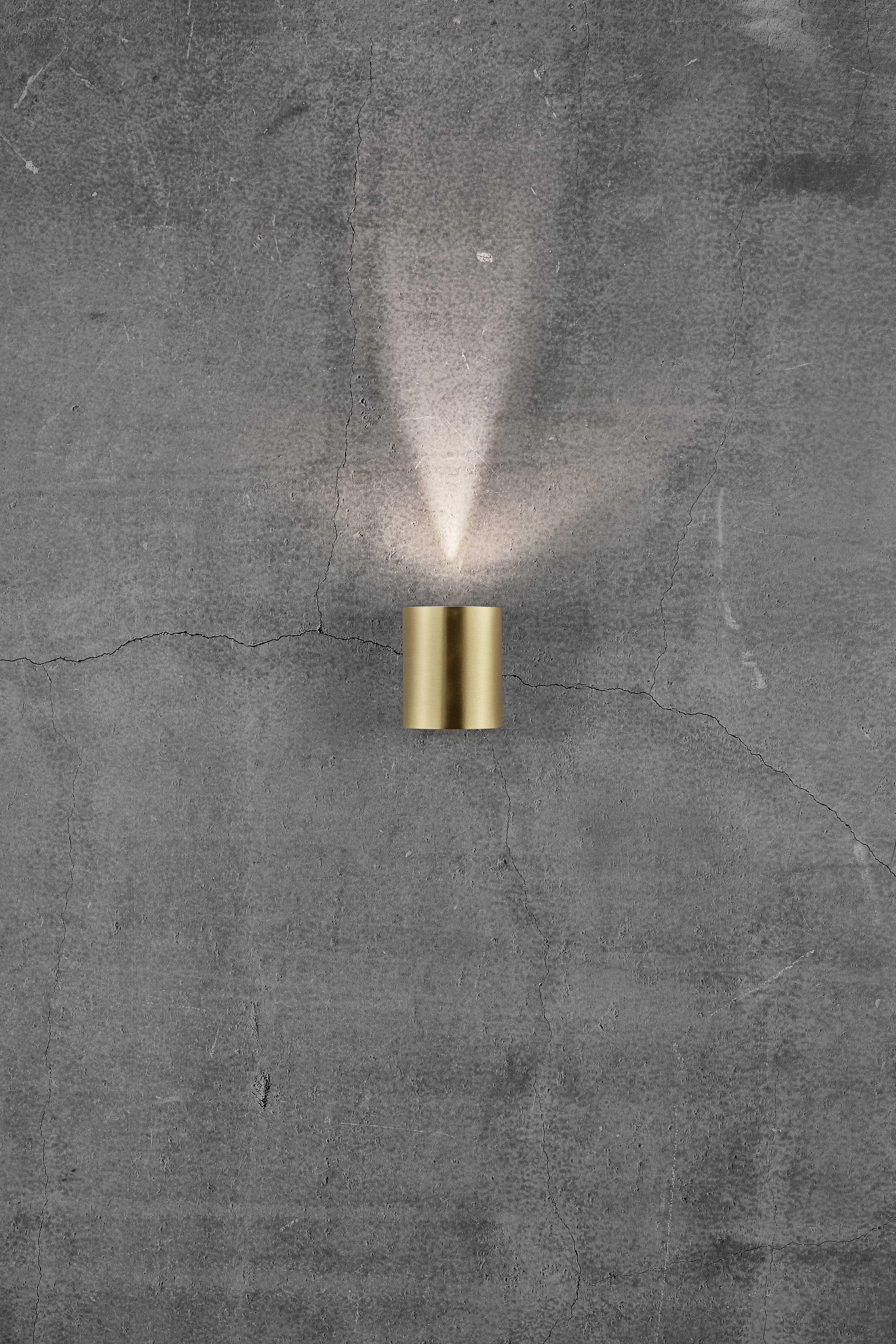 Wall light with a cylindrical, golden body, radiating warm light upwards and downwards, mounted on a grey concrete wall.