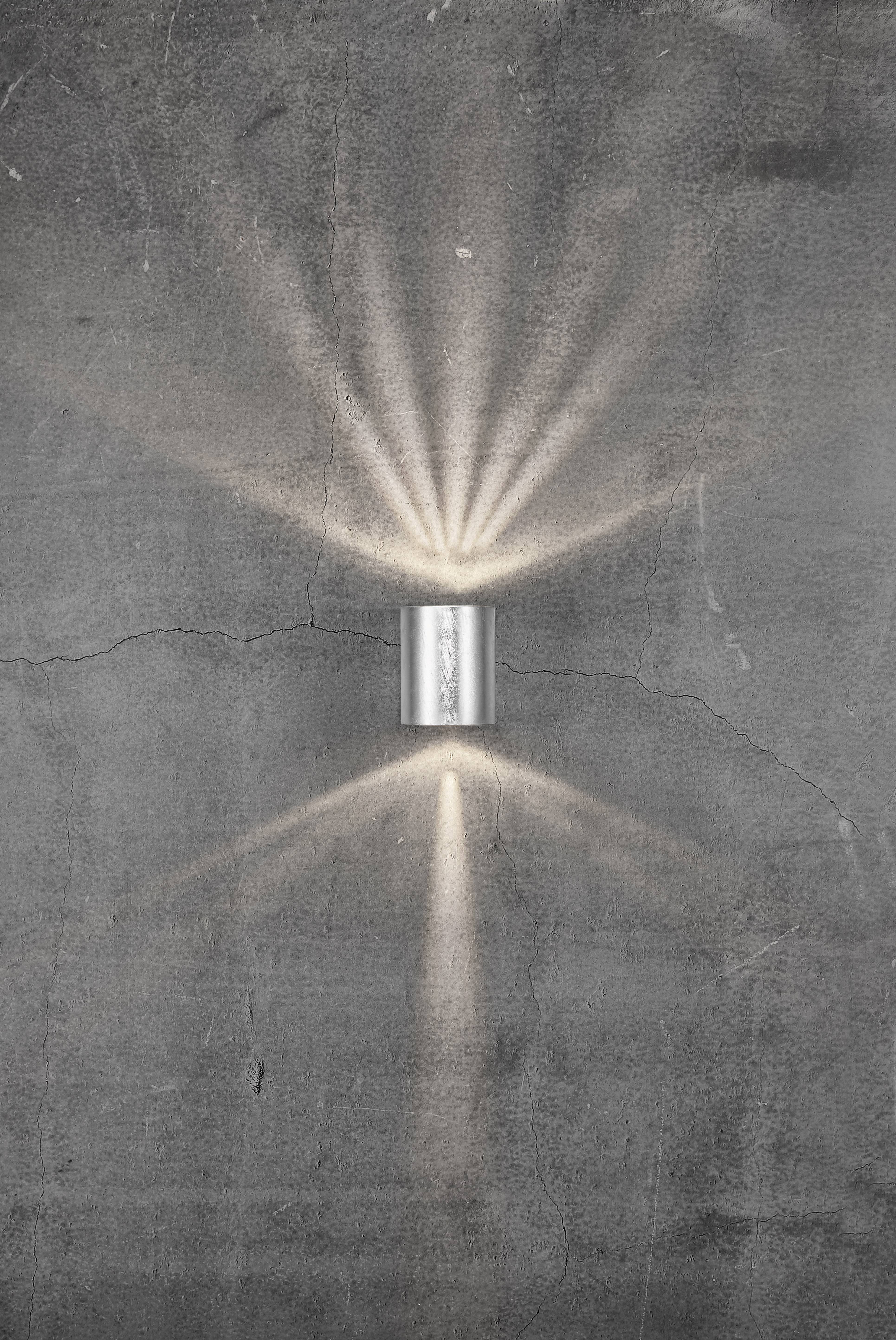 Silver wall light on a grey, cracked wall, radiating warm light upwards and downwards, creating soft light patterns.