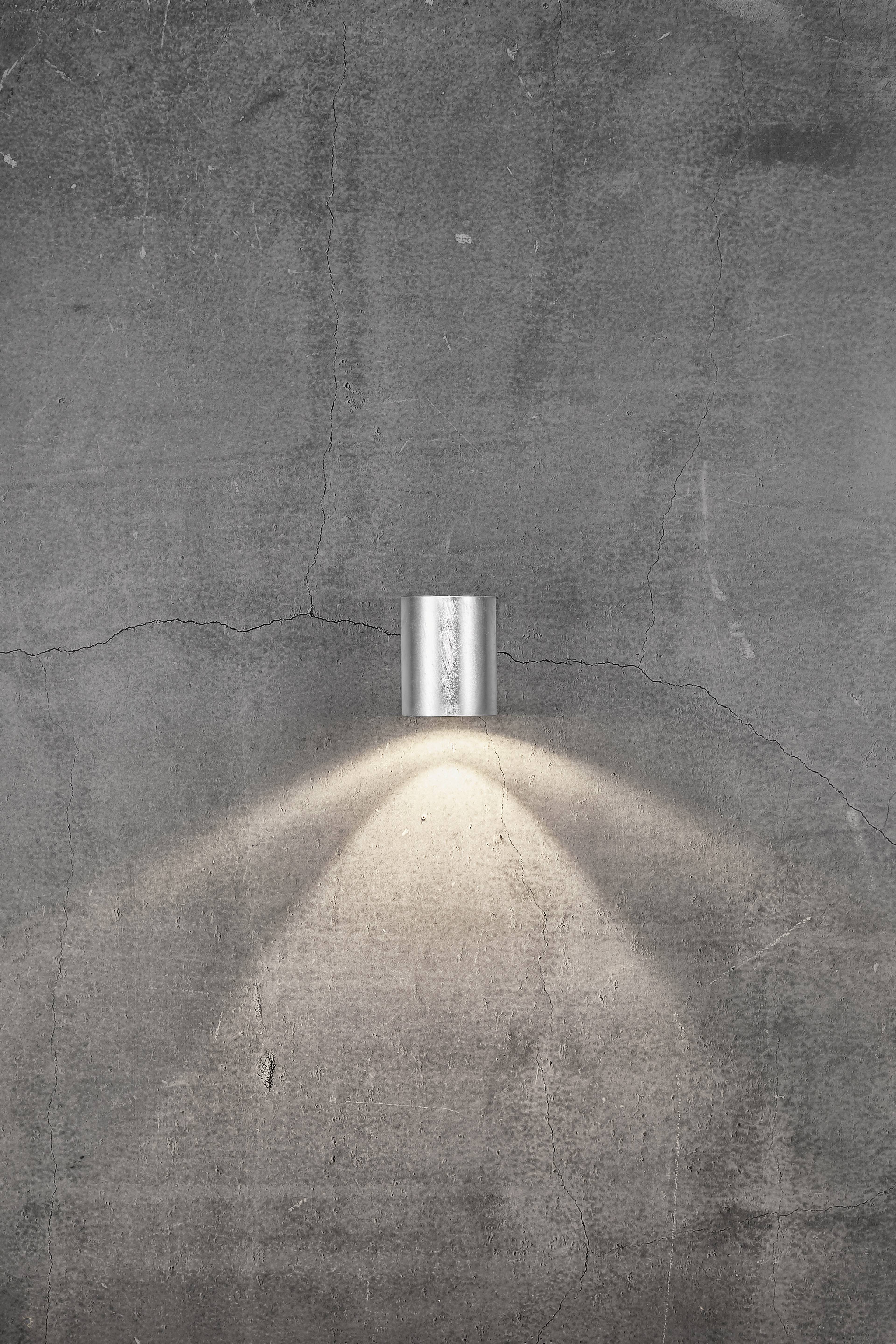 A silver wall light beams downwards, illuminating a dark, grey concrete wall and creating a wide cone of light.