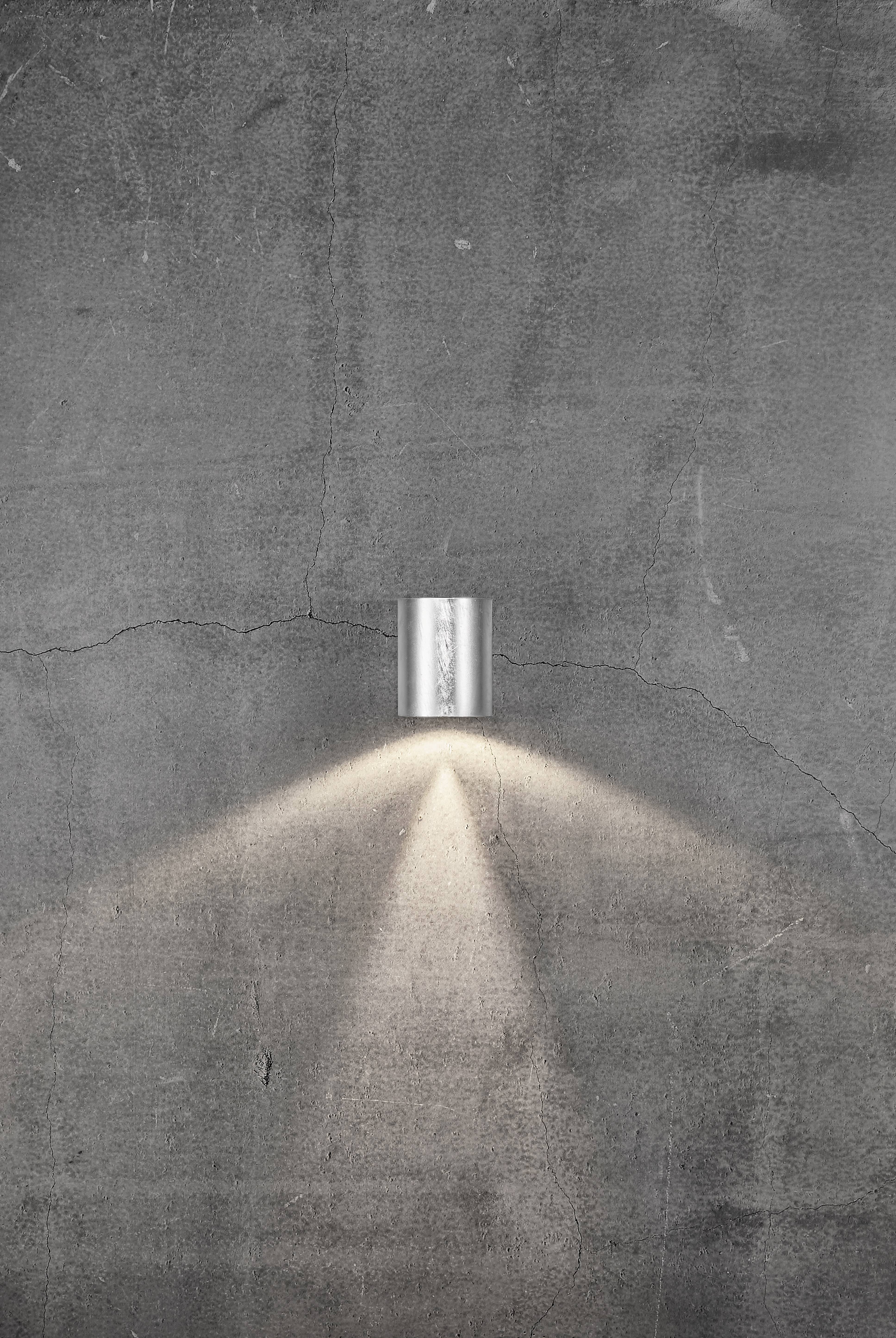A contemporary wall light on a rough, grey concrete wall, casting downward warm light, forms a V-shaped light cone.