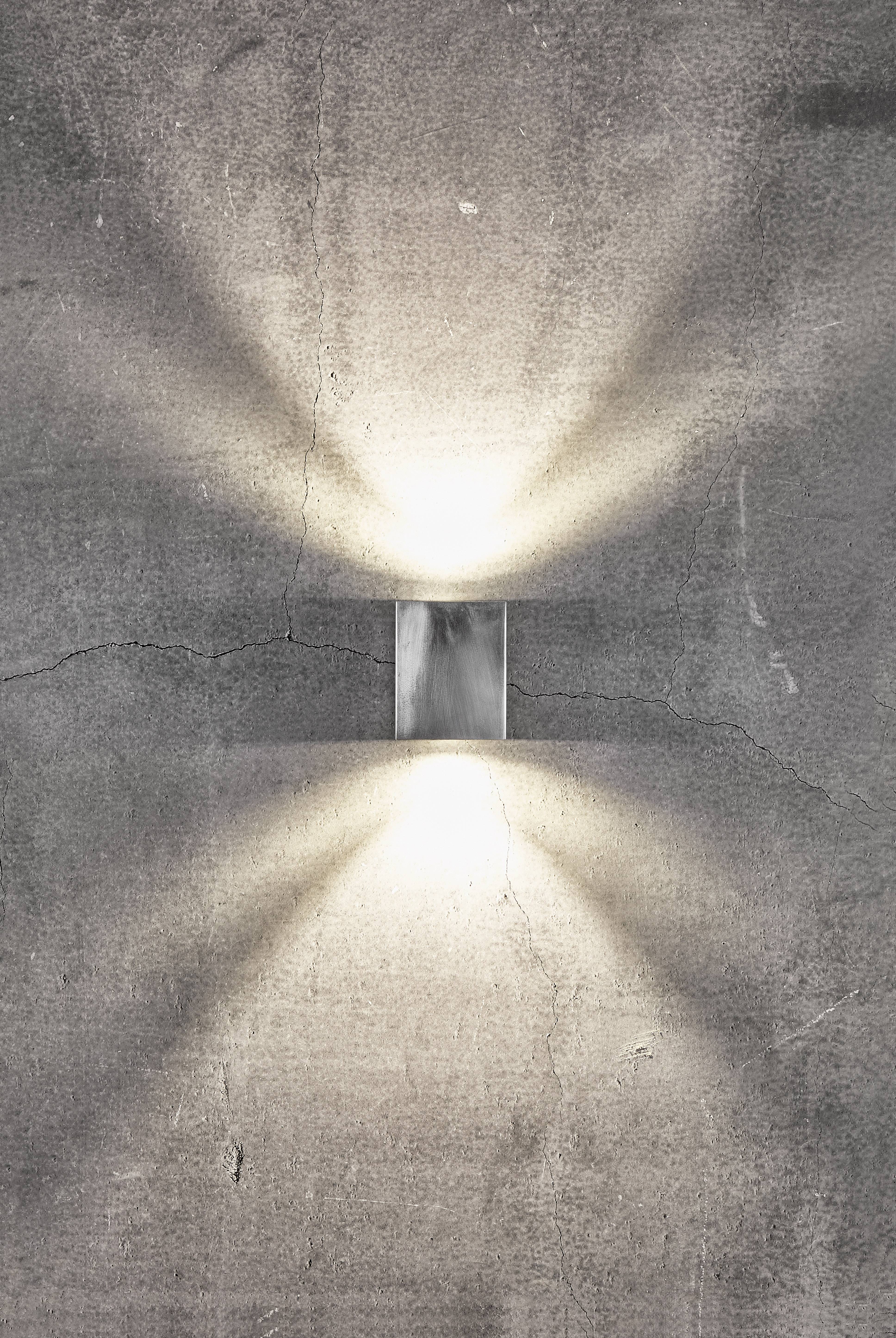 Wall light mounted on a grey concrete wall shines upwards and downwards, creating radiant light patterns on the wall surface.