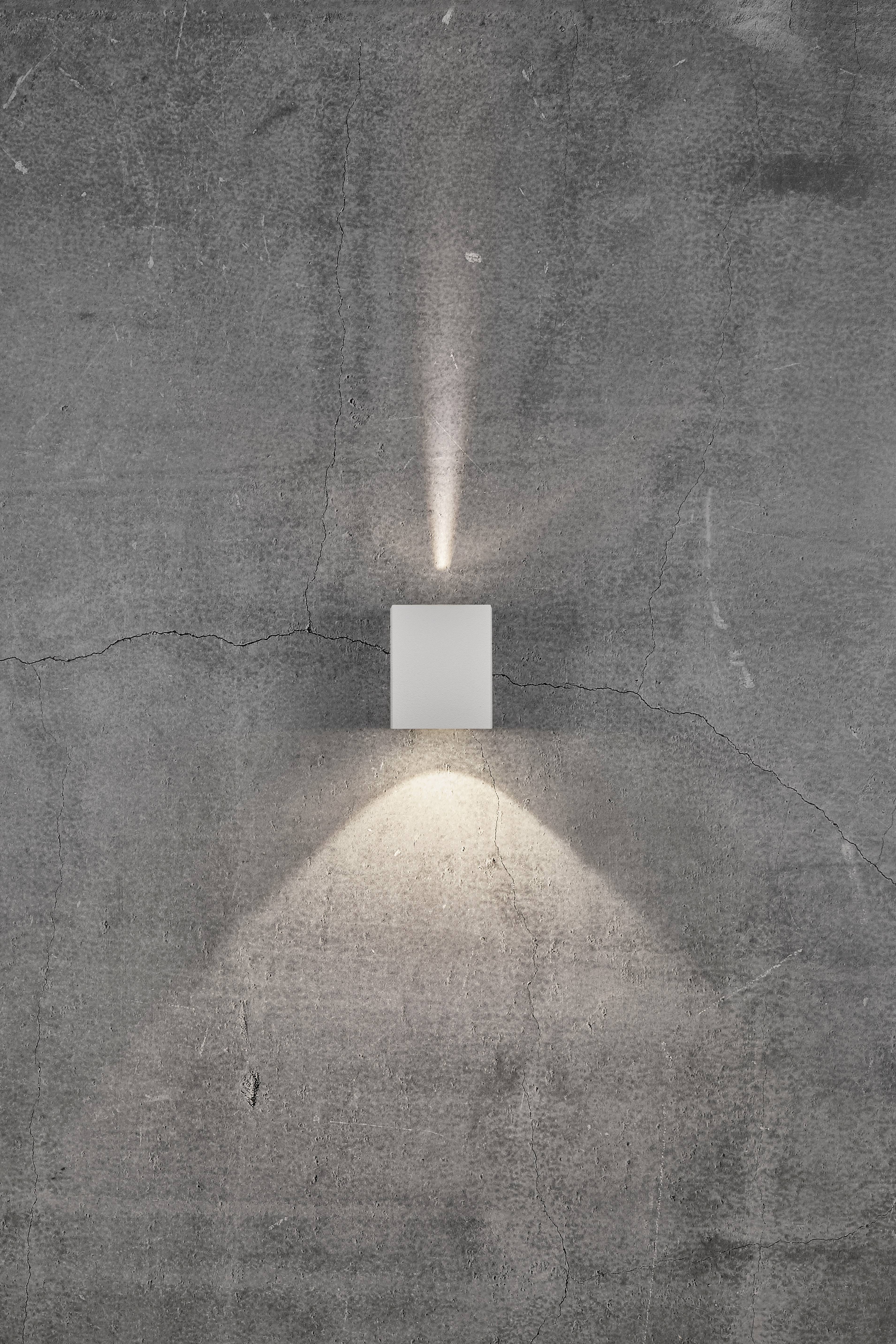 A minimalist wall light casts light upwards and downwards against a grey concrete wall, highlighting the texture of the surface.