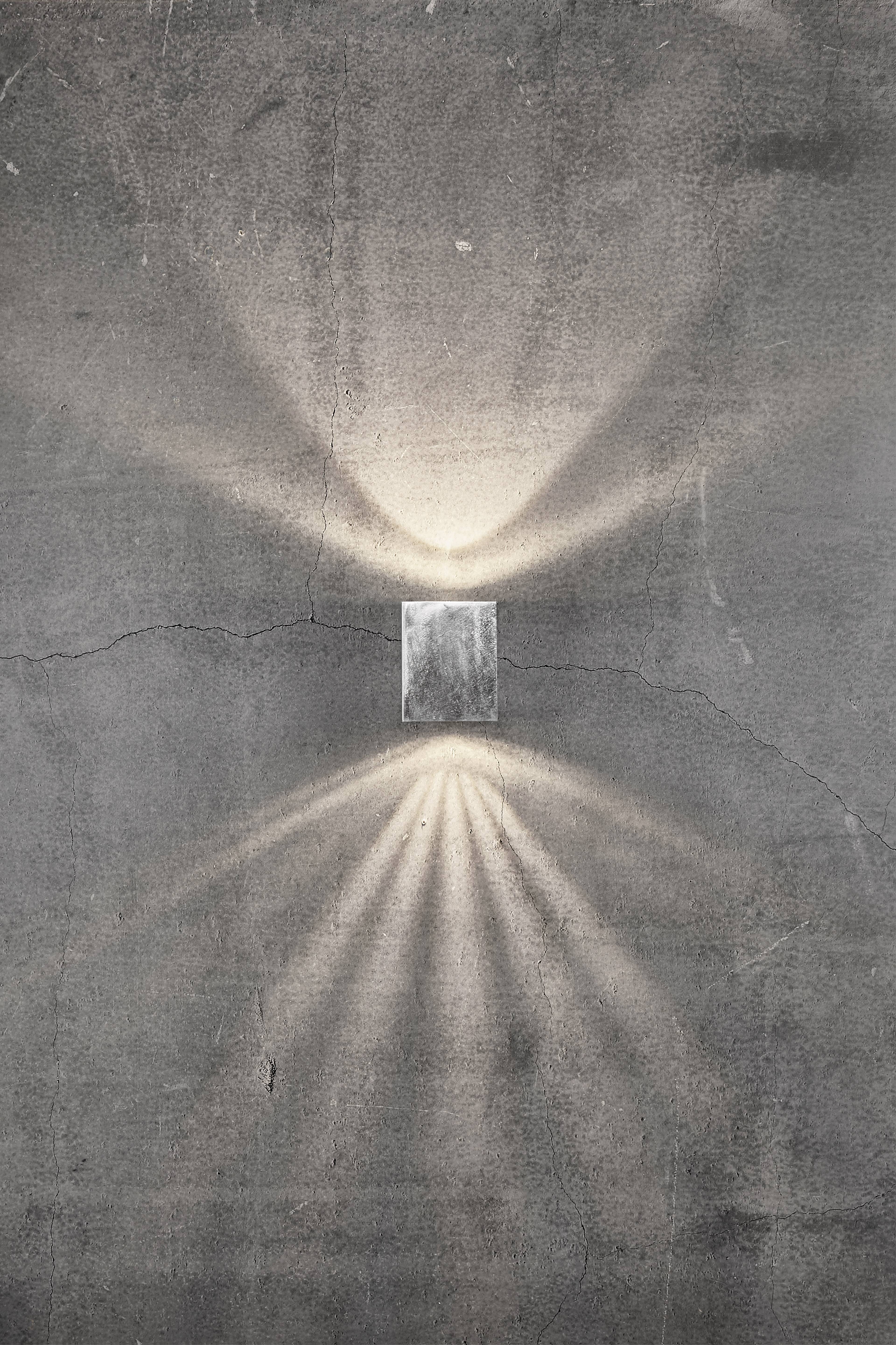 A rectangular wall light on a grey, textured wall. The light radiates upwards and downwards, creating decorative patterns.