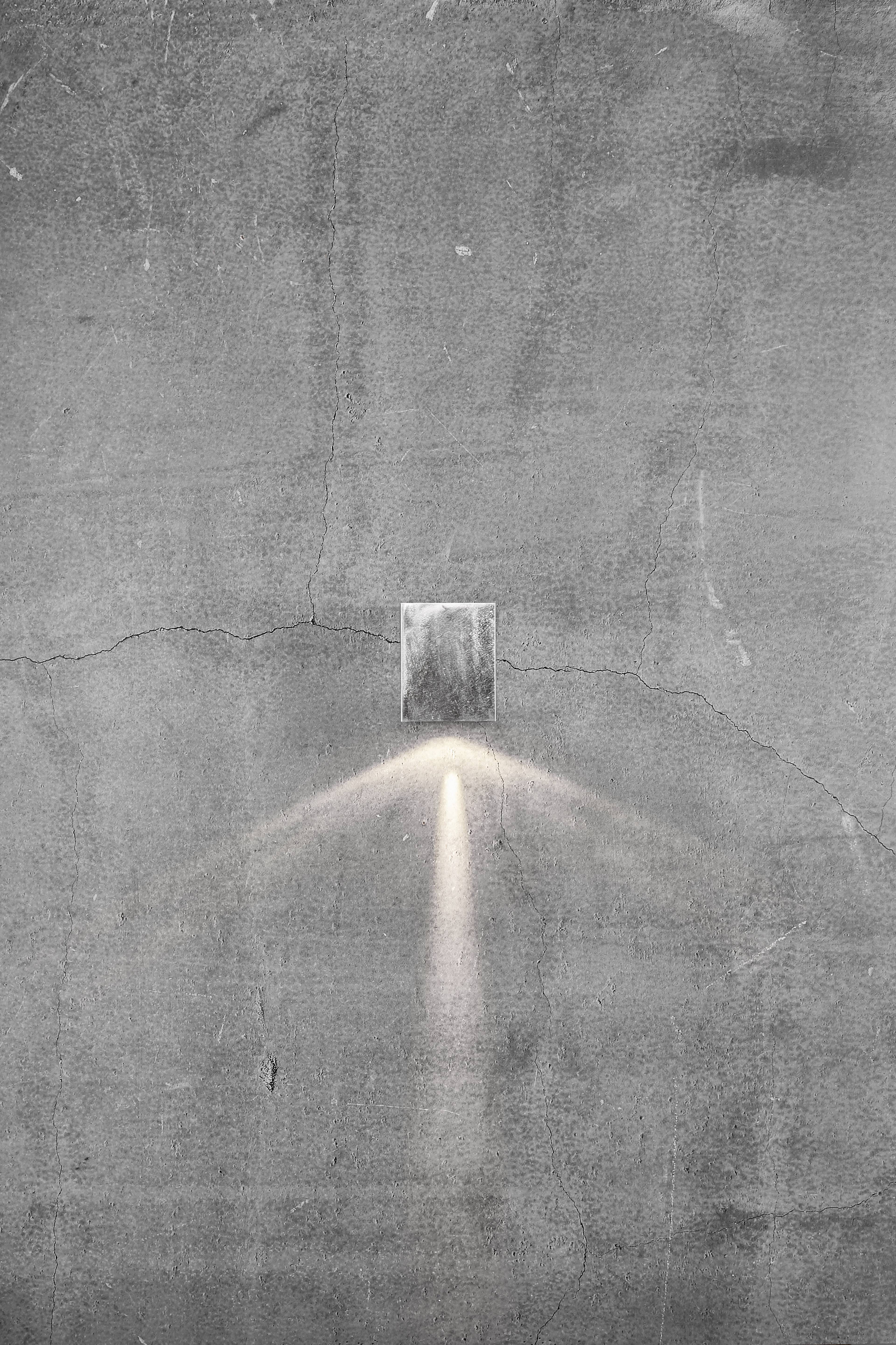 A rough concrete wall with a small rectangular light inlet, from which two light rays shine downwards.