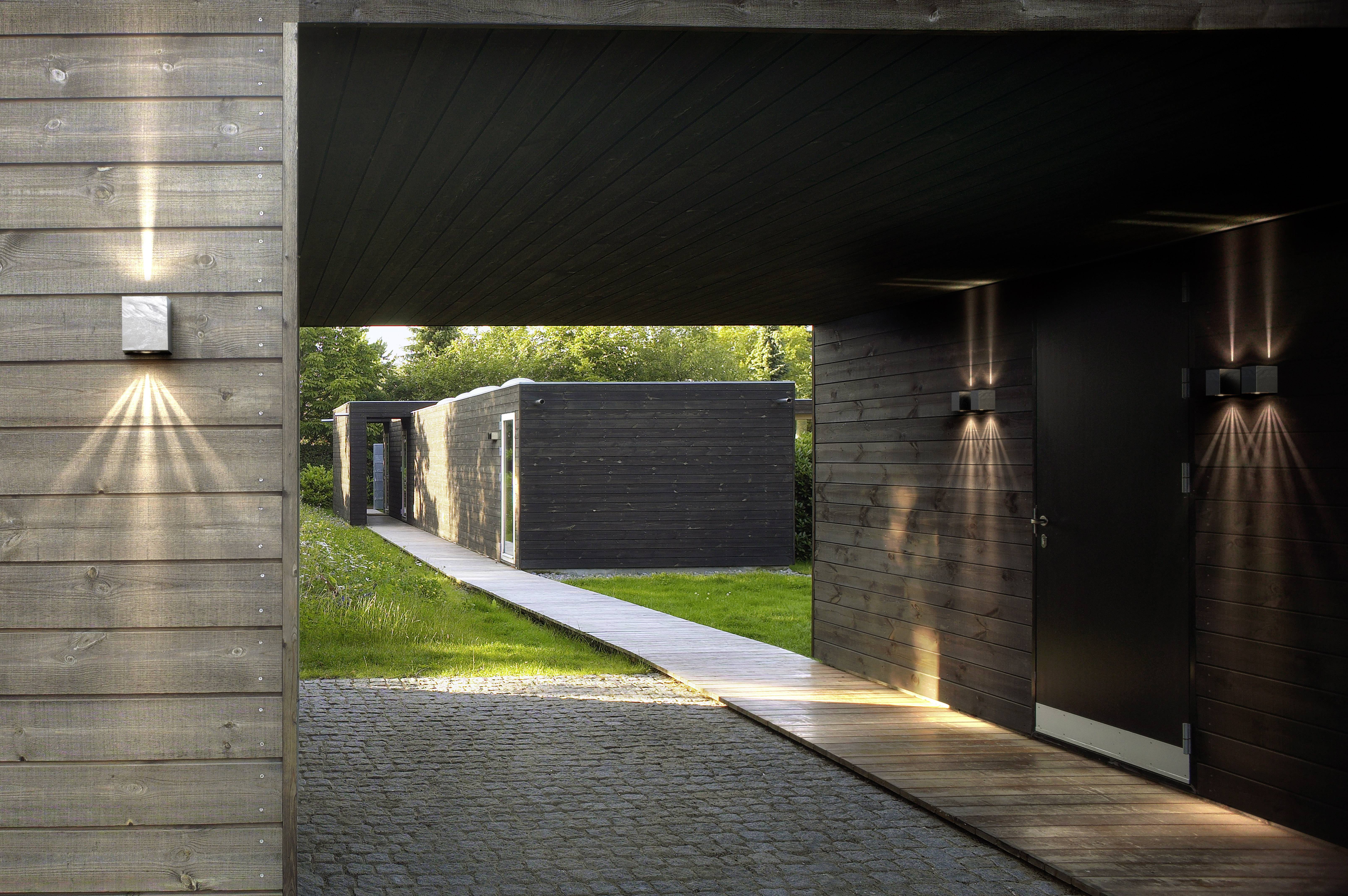 A modern timber-framed building complex with rectangular structures. A paved pathway leads through a covered area into the greenery.