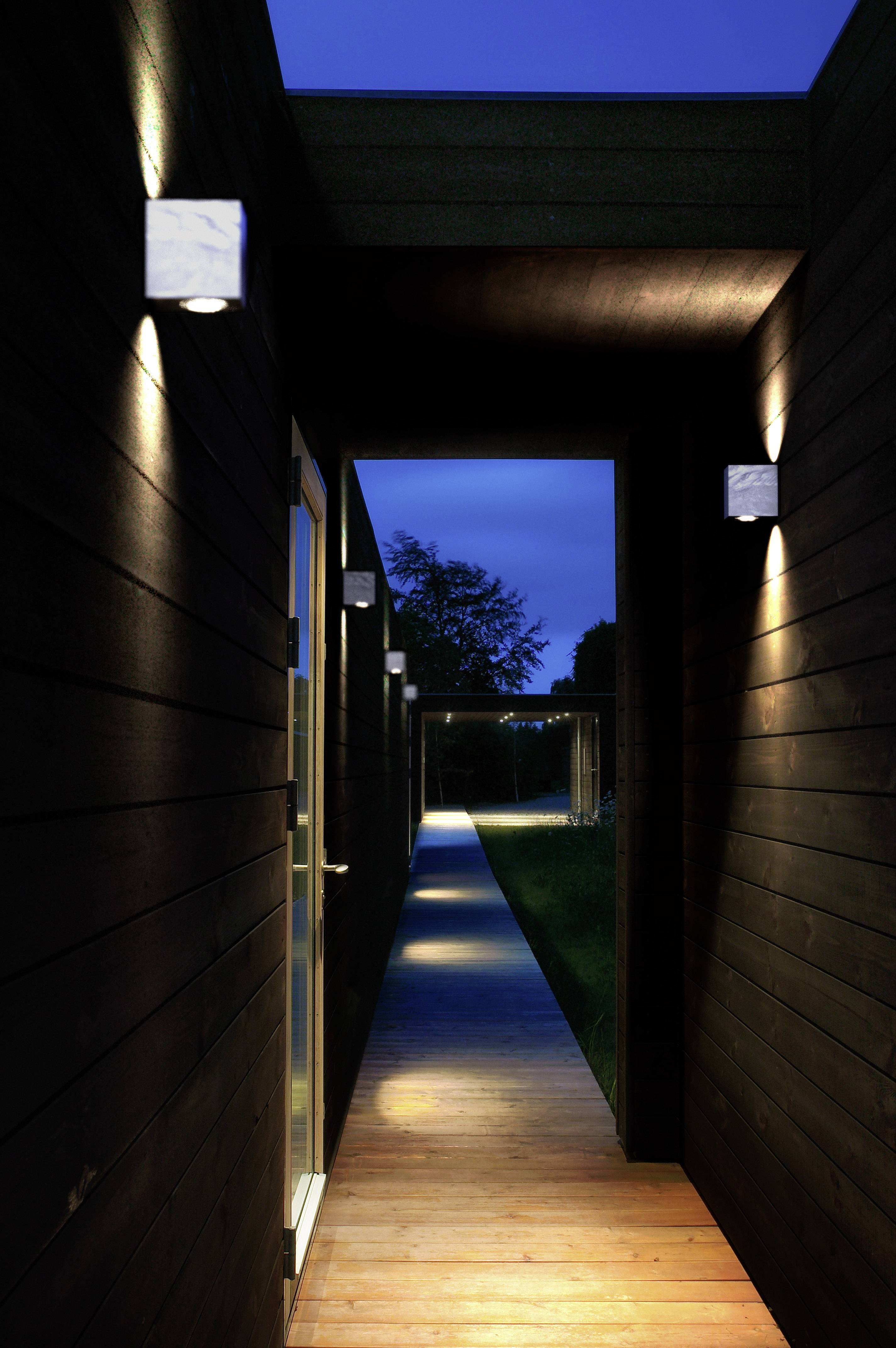 A narrow, illuminated wooden passageway with lamps on the walls leads to a lit outdoor area, accompanied by a dark sky.