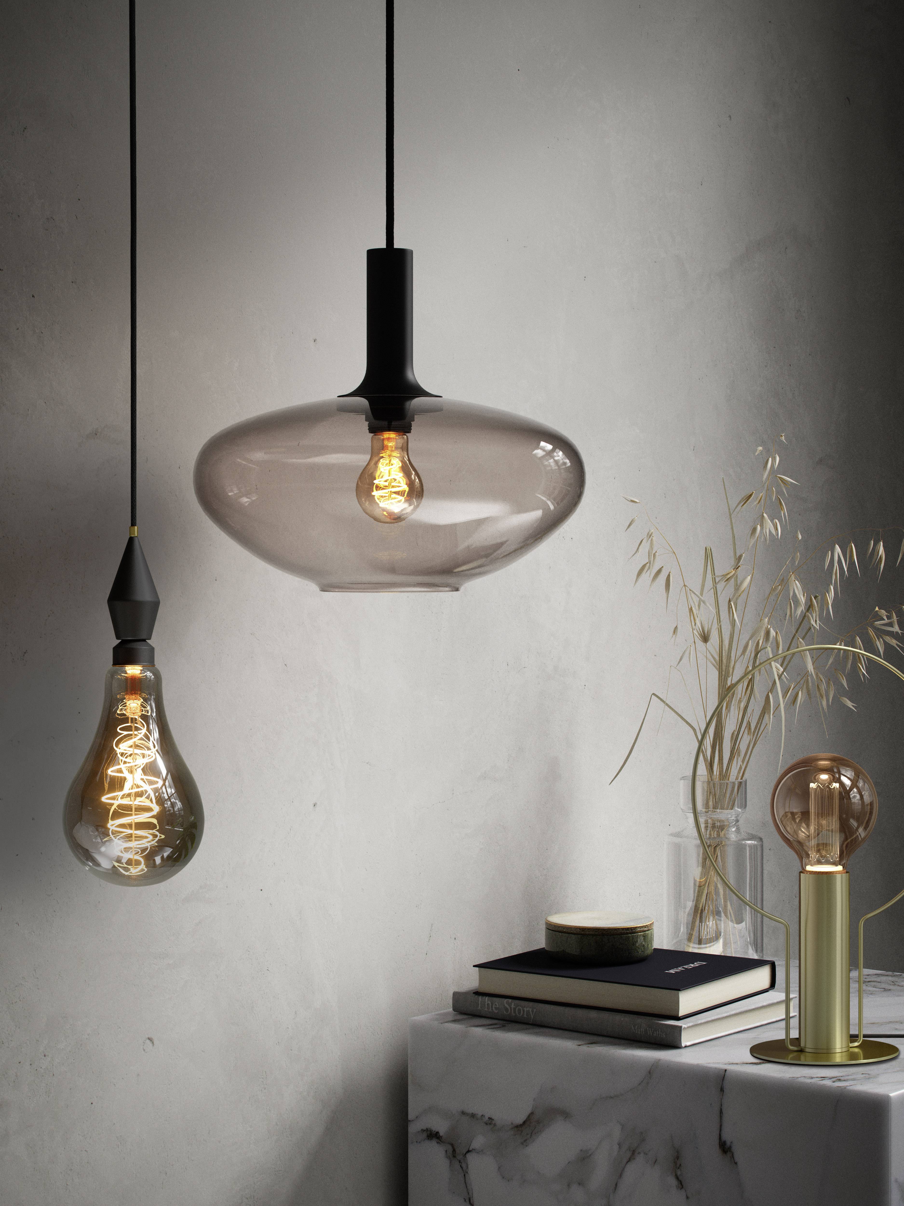 Two hanging retro light bulbs in front of a grey wall, beside a table with books, a small vase, and a glowing table lamp.