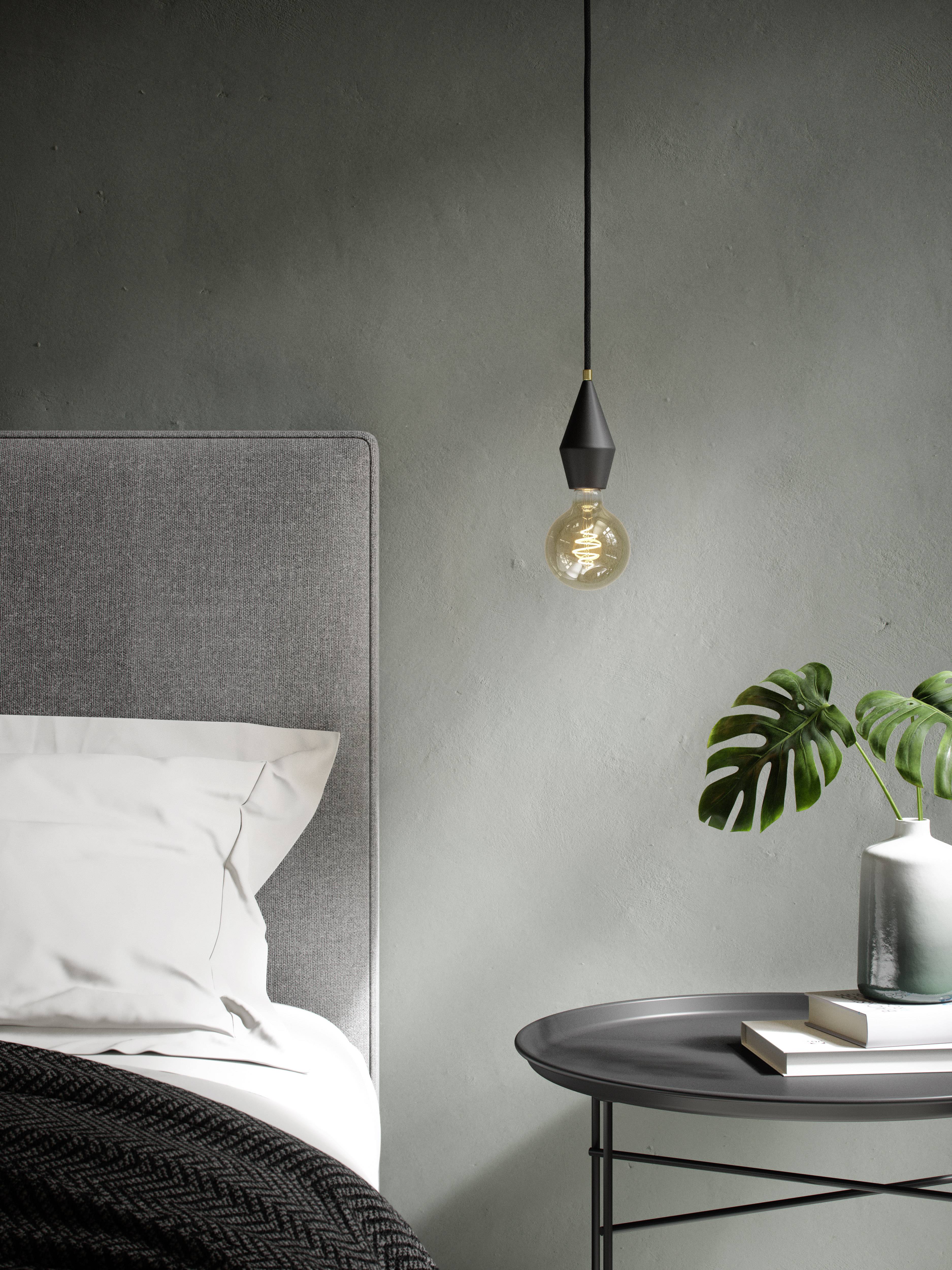 A grey bedroom with a bed, a hanging light bulb, and a table with a green plant and a candle on it.