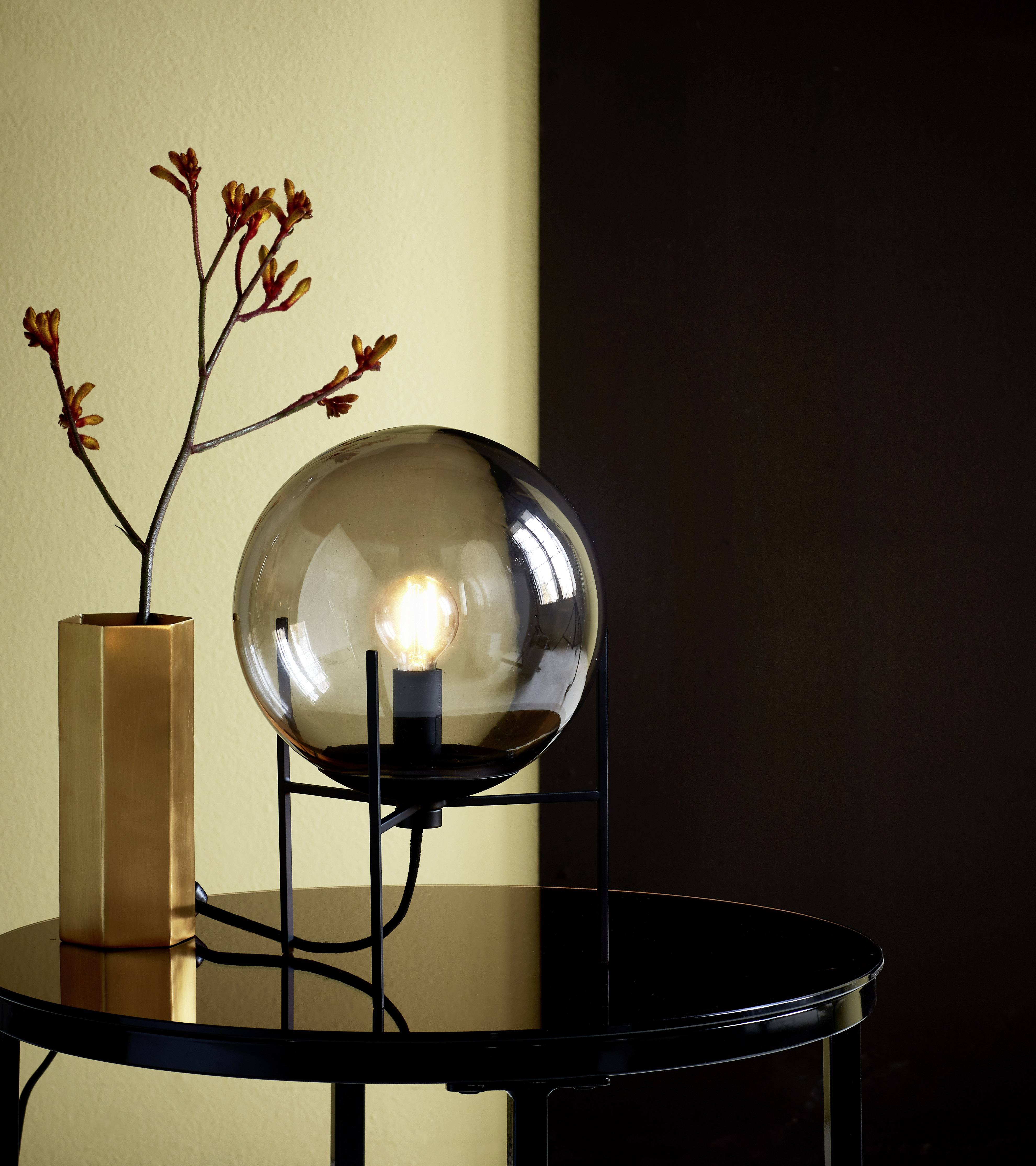 Round table lamp with smoky grey glass and black frame on a table. Next to it stands a vase with branches against a dark background.