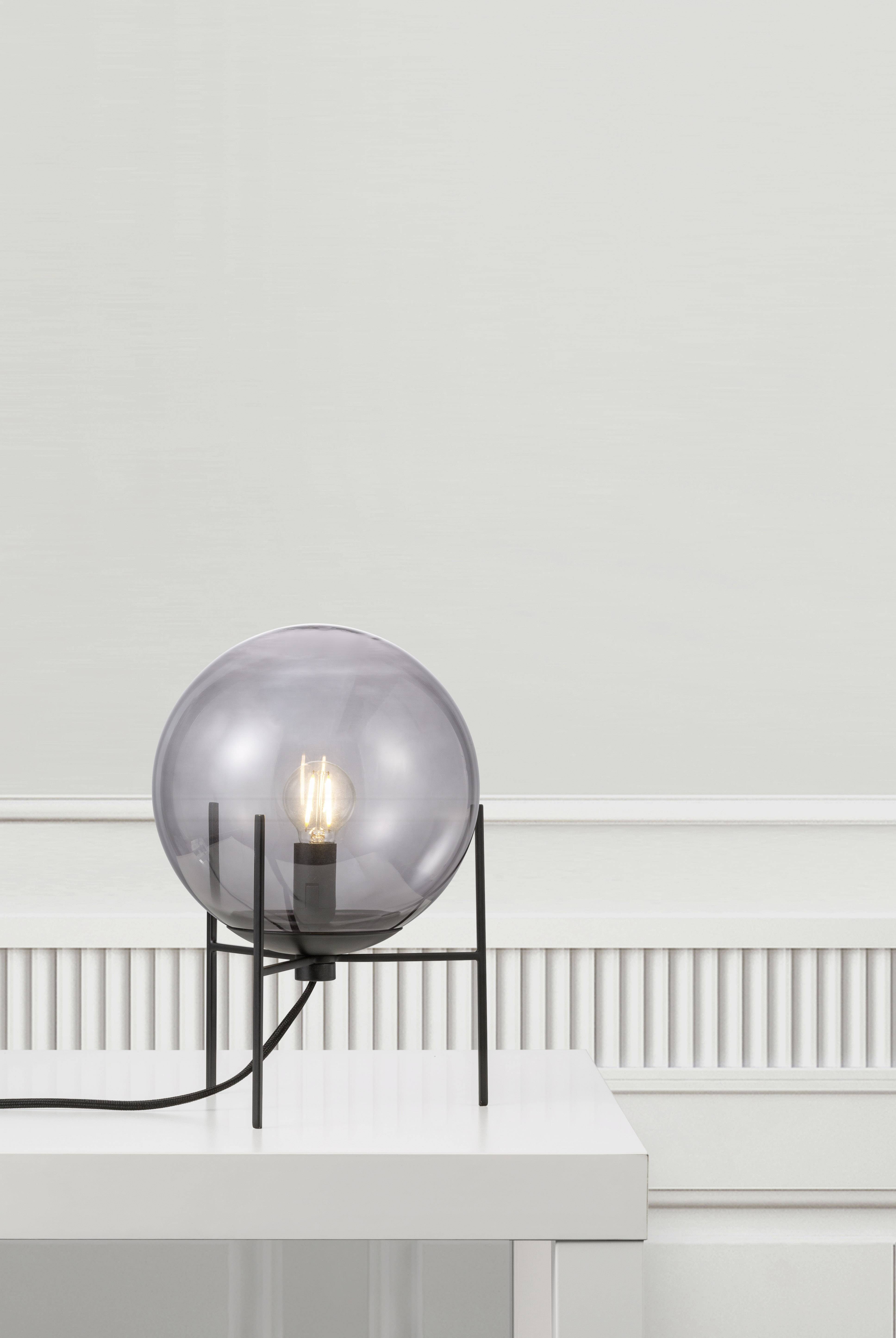 A modern table lamp with a spherical, transparent shade stands on a white table. It emits warm light.