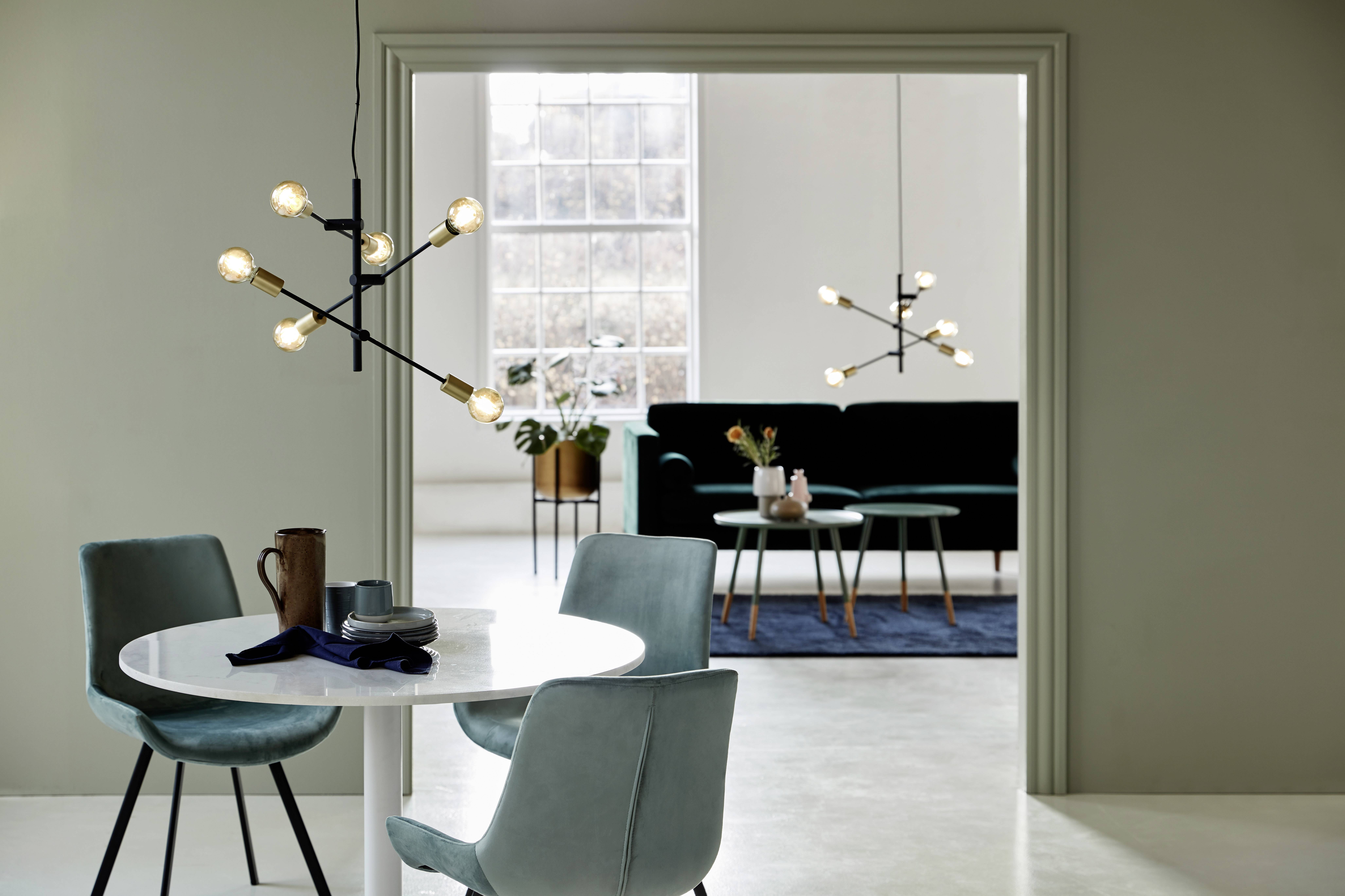 Dining room with a round table and four chairs, with a living room in the background featuring a sofa and rug. Modern chandeliers illuminate both spaces.