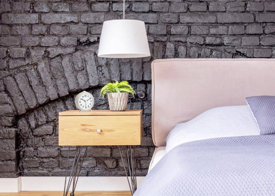 A modern bedroom with a floor lamp positioned above a wooden bedside table. A clock and a small plant sit atop the table. Background: brick wall.