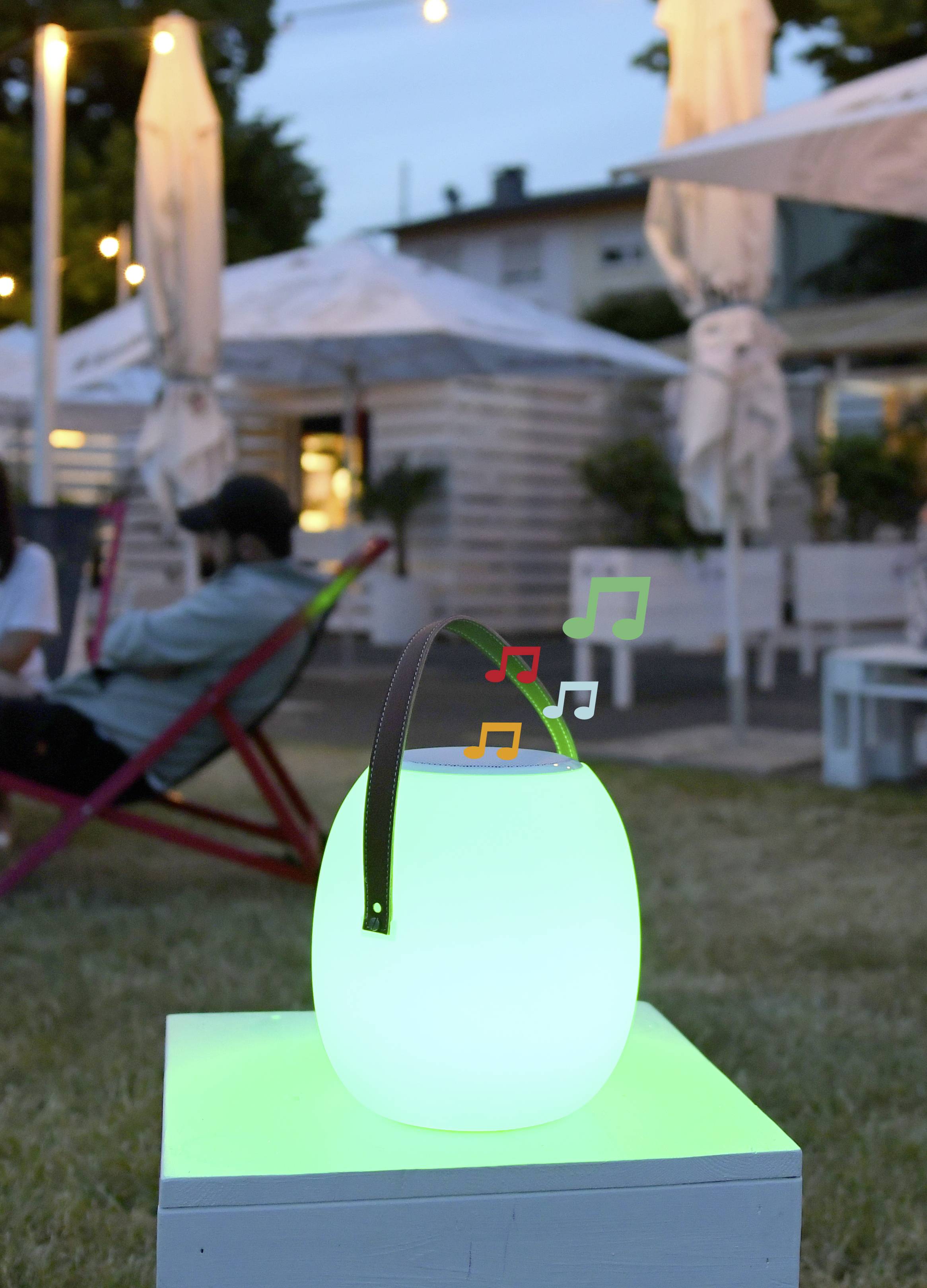 A luminous lamp with musical notes sits in the foreground on an outdoor table. In the background, people are seated on loungers.