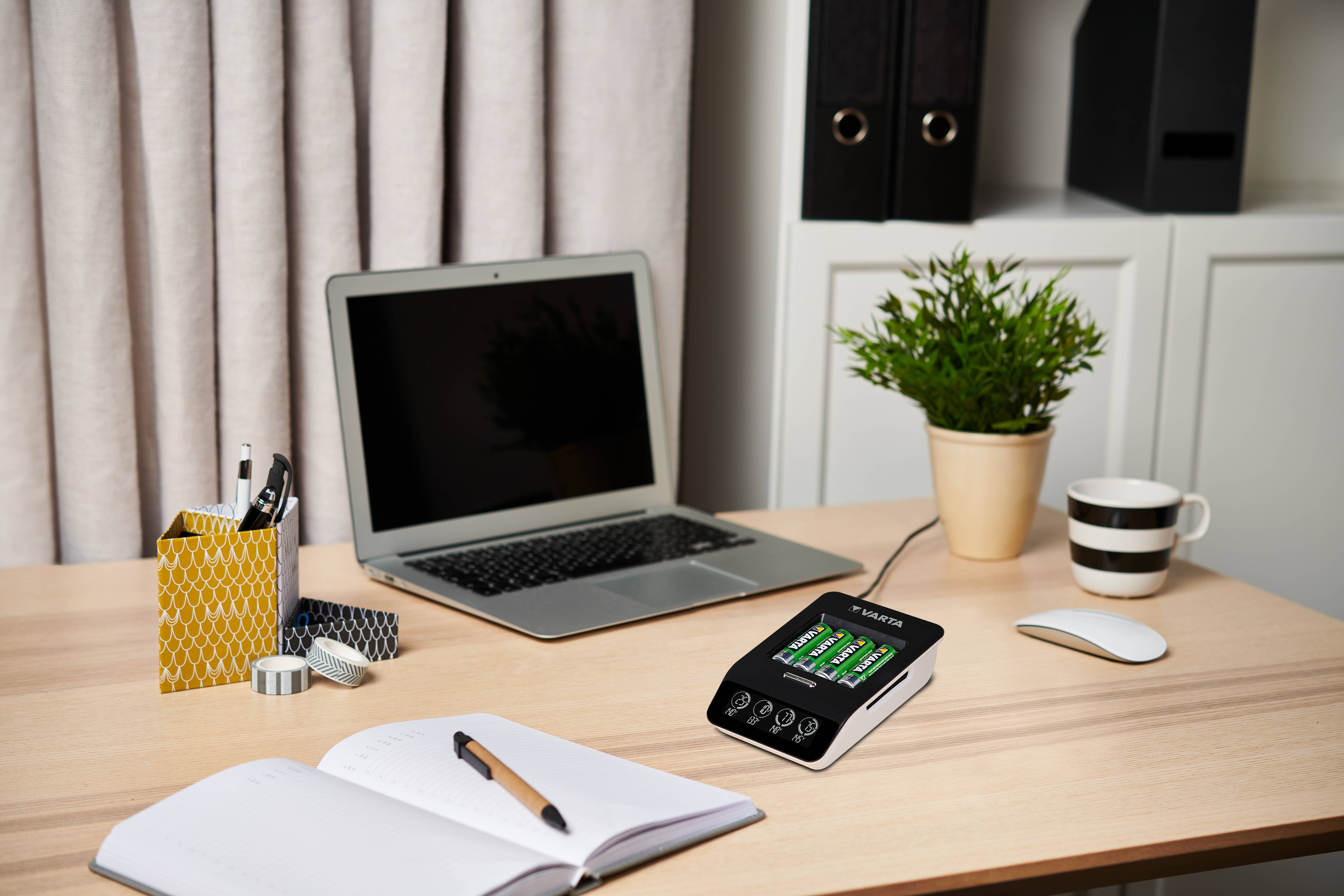 A tidy desk with a laptop, open notebook, plant, and a charging device displaying battery levels. A cup and office supplies are present.