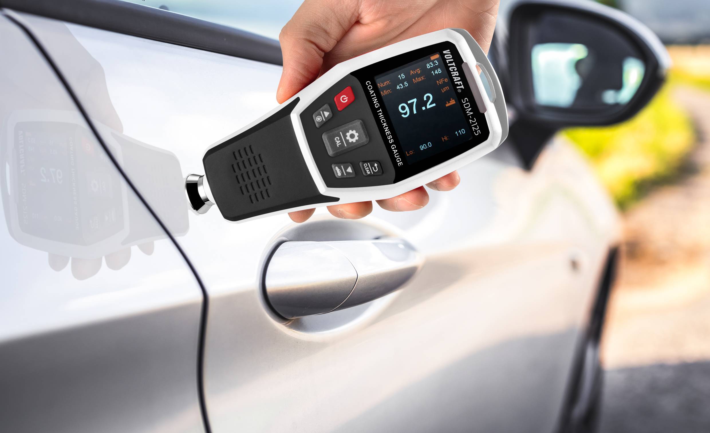 A person is holding a paint thickness gauge against a car door, with the display showing '97.2'. The car is light grey, with a blurry background.