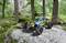A remote-controlled car with a colourful pattern sits on a large rock in the woods, surrounded by tall trees and green moss.