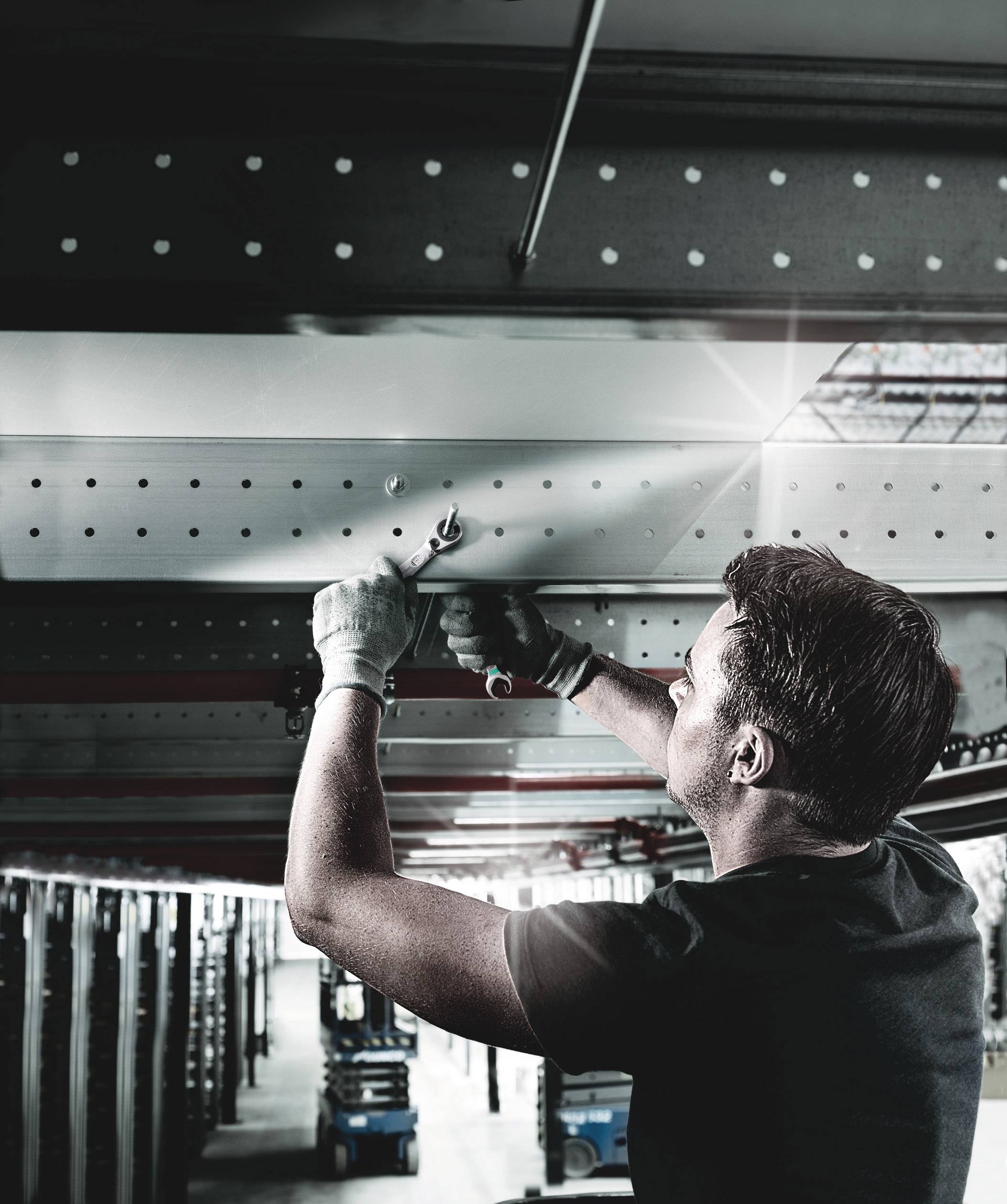 A man is installing metal beams beneath a roof in an industrial setting, focused and with tools in his hands.