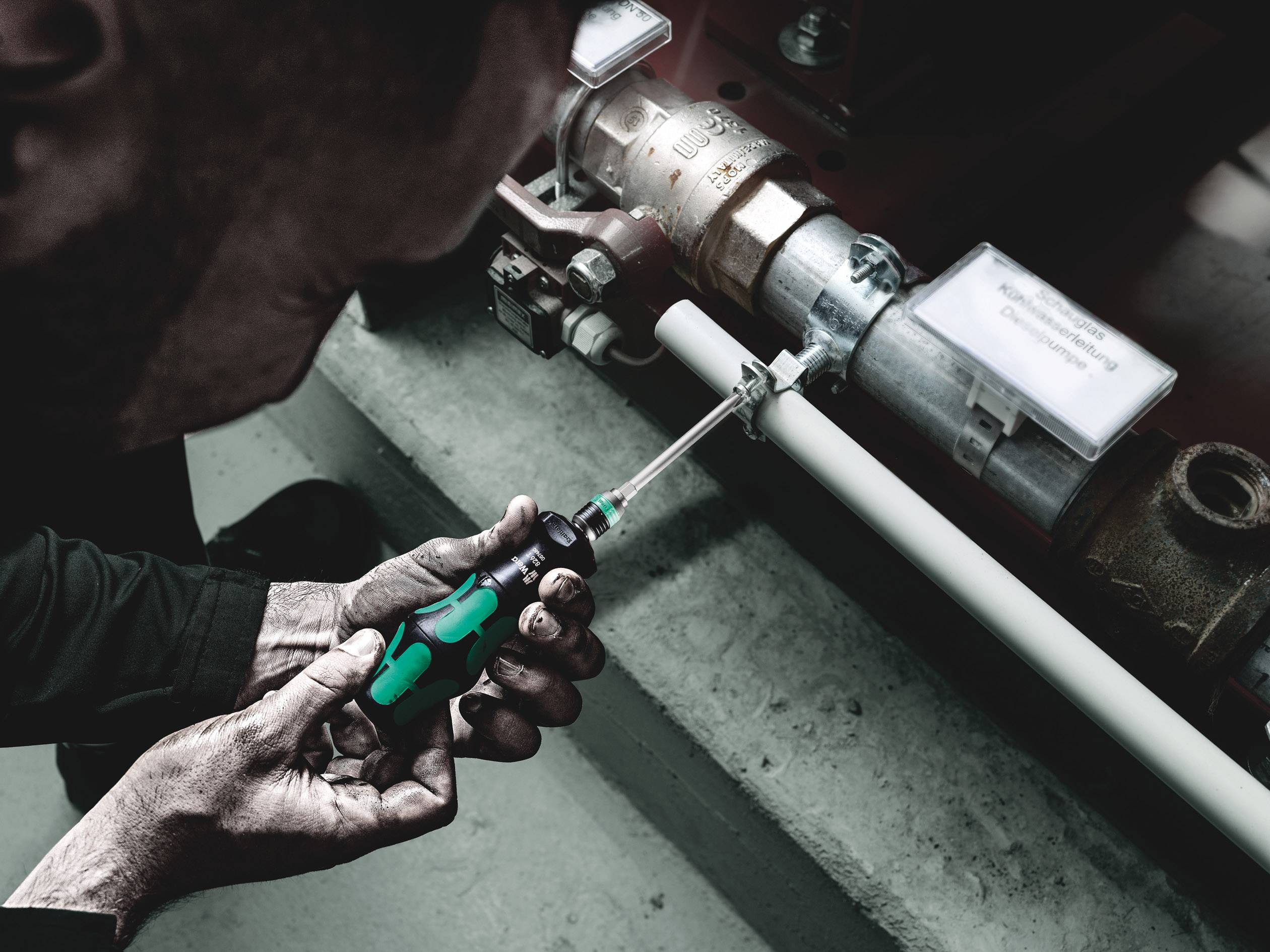 A person is tightening a screw on a pipe system with a green screwdriver, in an industrial setting.