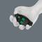 A white hand is holding a small black device with green symbols, resembling a controller, against a grey background.