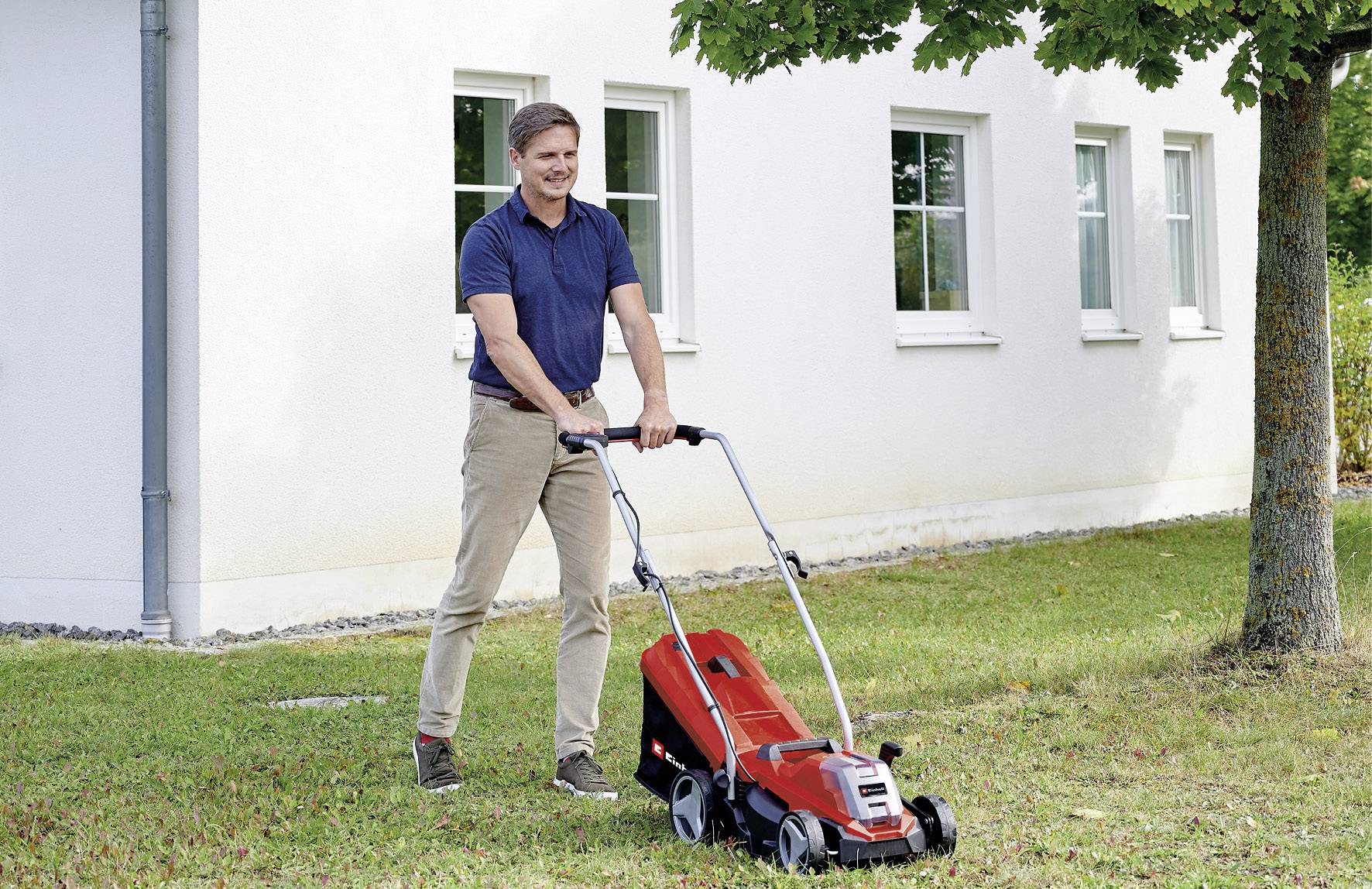 A man is mowing the lawn in front of a white house with a red lawnmower. A tree stands to the right of the house.