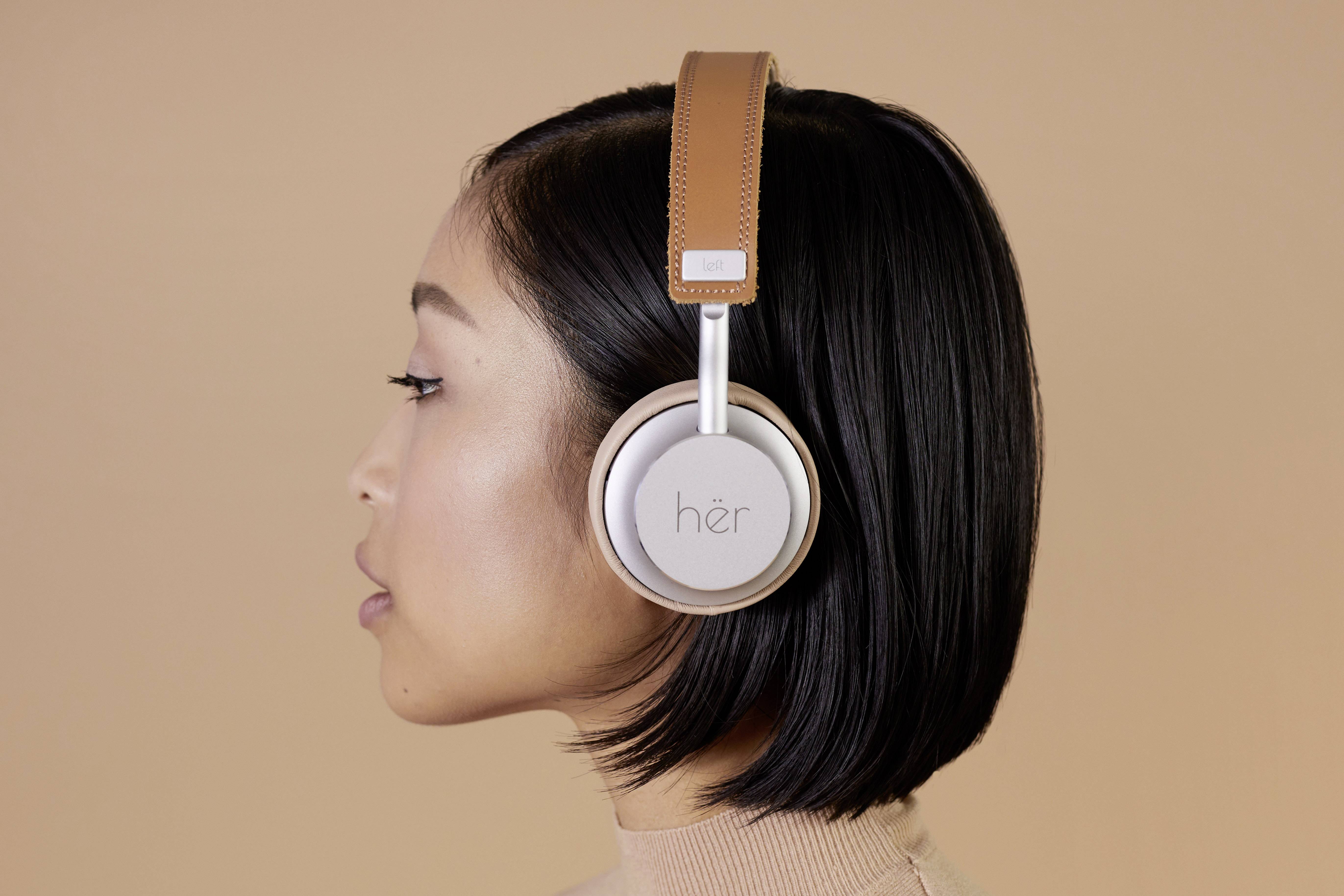 A person with sleek, dark hair is wearing large, brown headphones against a plain background.