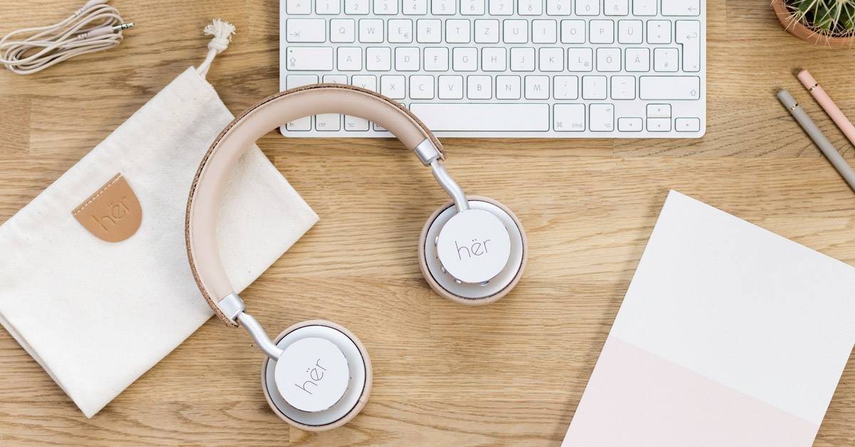Desk with keyboard, headphones, notebook and pens on a wooden surface. White and pink headphones are the central element.