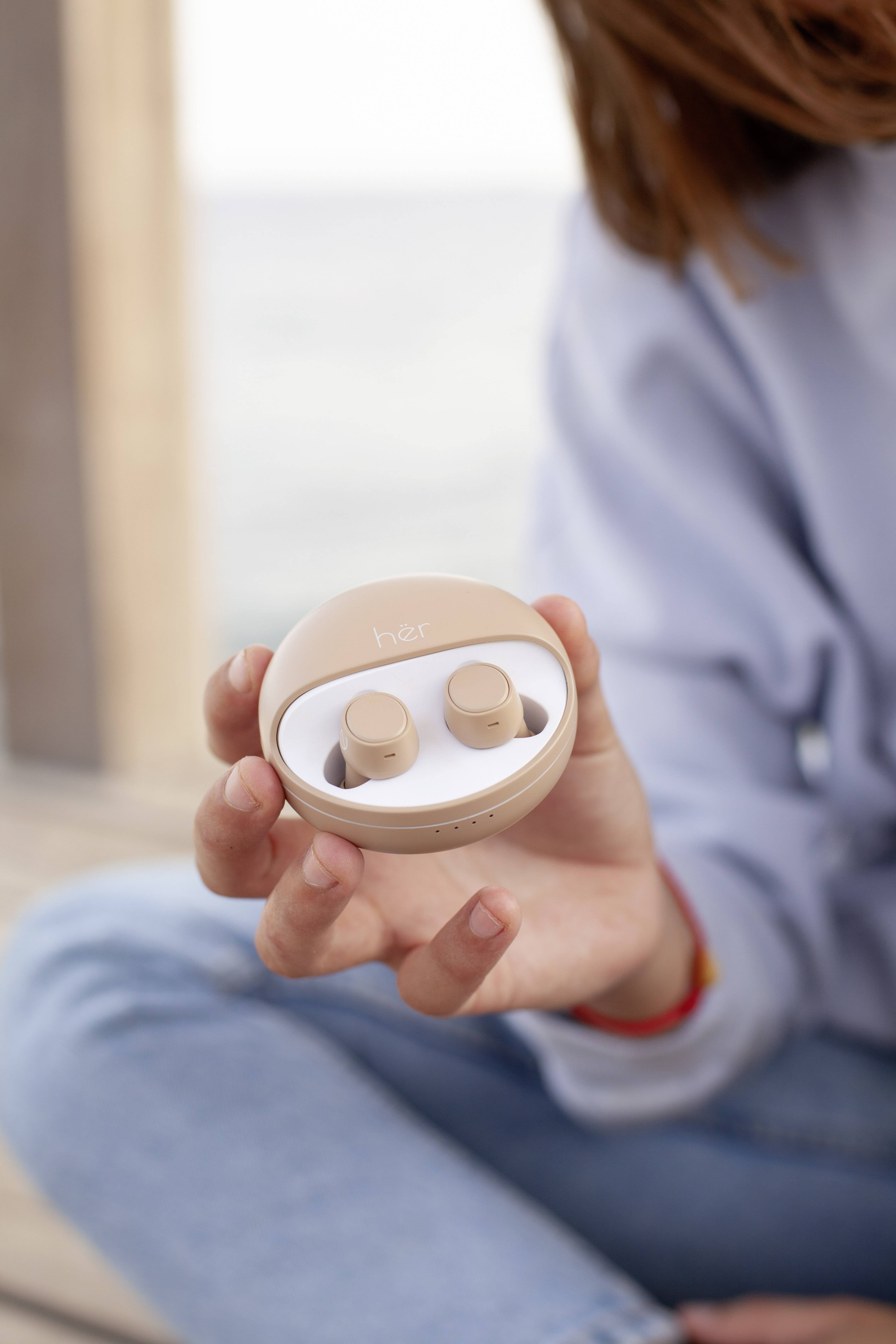 A person is holding a round box containing wireless earbuds, with beige-coloured earbuds visible inside the box. The background is blurred.